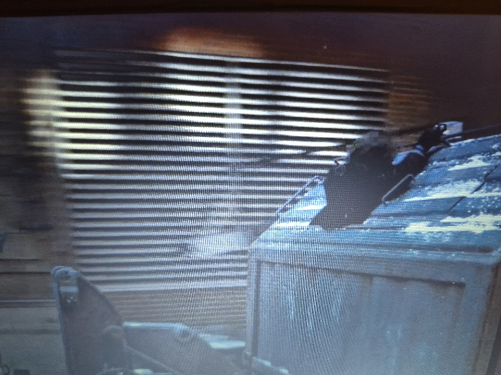 The image depicts a distressed and aged industrial scene, possibly in Sandwell, United Kingdom. A dark, bird-like silhouette, potentially a crow or raven, perches atop a weathered, blue corrugated structure. The structure itself appears to be a container or shed, with visible signs of rust and decay. Behind it, a wall with horizontal slats or blinds is visible, casting striped shadows. The overall atmosphere is bleak and somewhat ominous, suggesting an abandoned or neglected area. There are no people or discernible human activity in the frame.