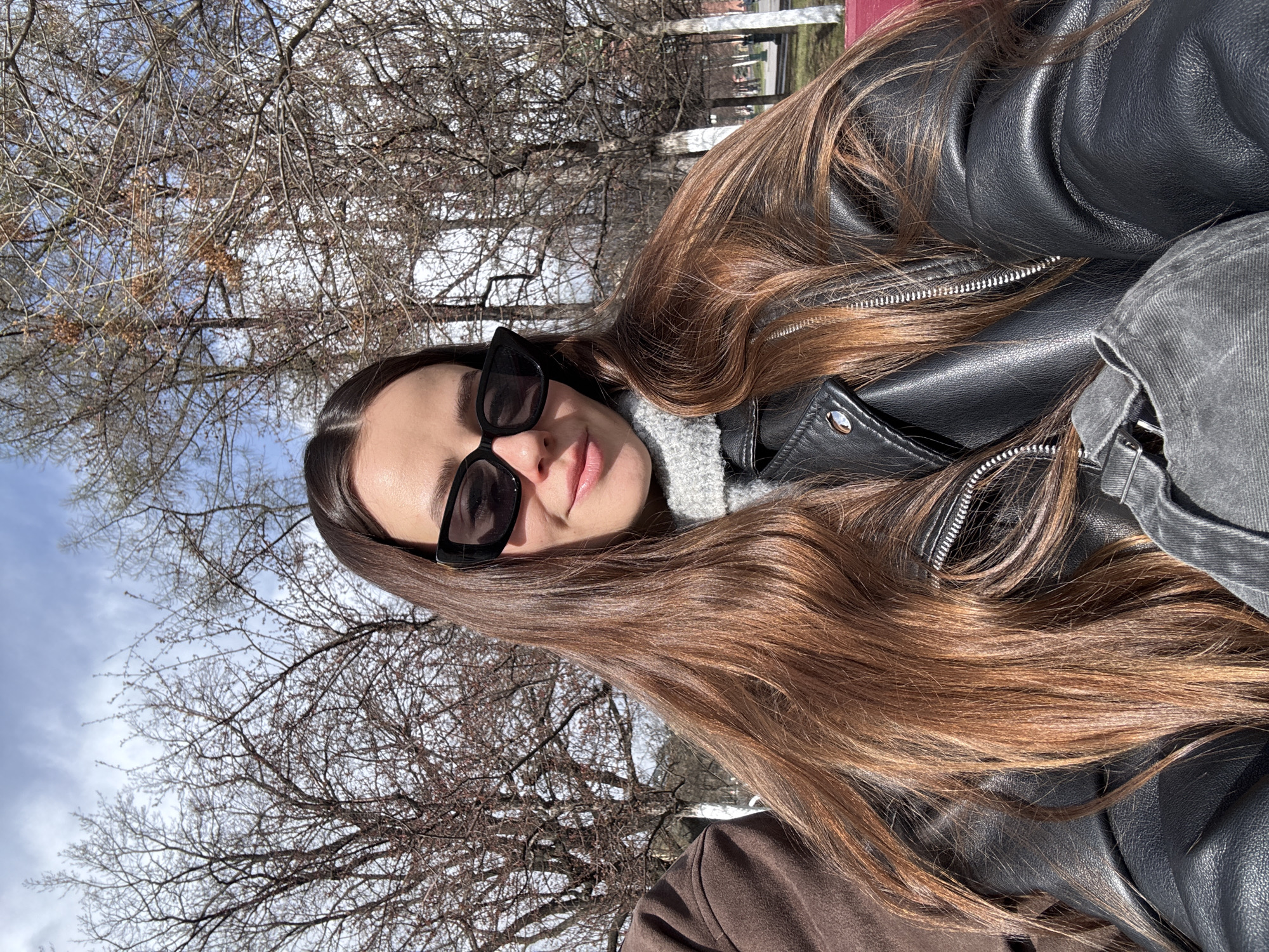 A woman with long brown hair, wearing sunglasses and a black leather jacket over a gray turtleneck, poses for a selfie. The background consists of bare trees and a partly cloudy sky. Given the architecture visible in the far background and the general ambiance, the location is likely a park or open space in Moscow, Russia, during the late fall or early spring. The person is looking at the camera.