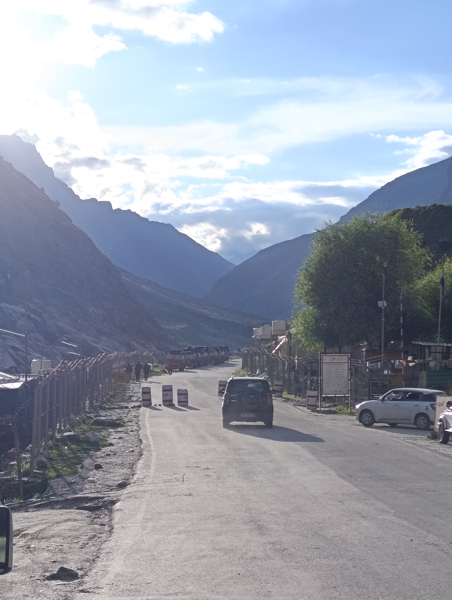 The image captures a roadside scene in Damphug, India, with towering mountains framing a clear sky and scattered clouds. A paved road runs through the center, with a line of parked trucks visible on the right side, suggesting a possible transport hub or resting area. Several vehicles, including a white SUV and a dark SUV, are present on the road. On the left, a chain-link fence separates the road from a rocky embankment with sparse vegetation. A few people are seen walking along the roadside. The overall environment appears to be mountainous and somewhat arid.