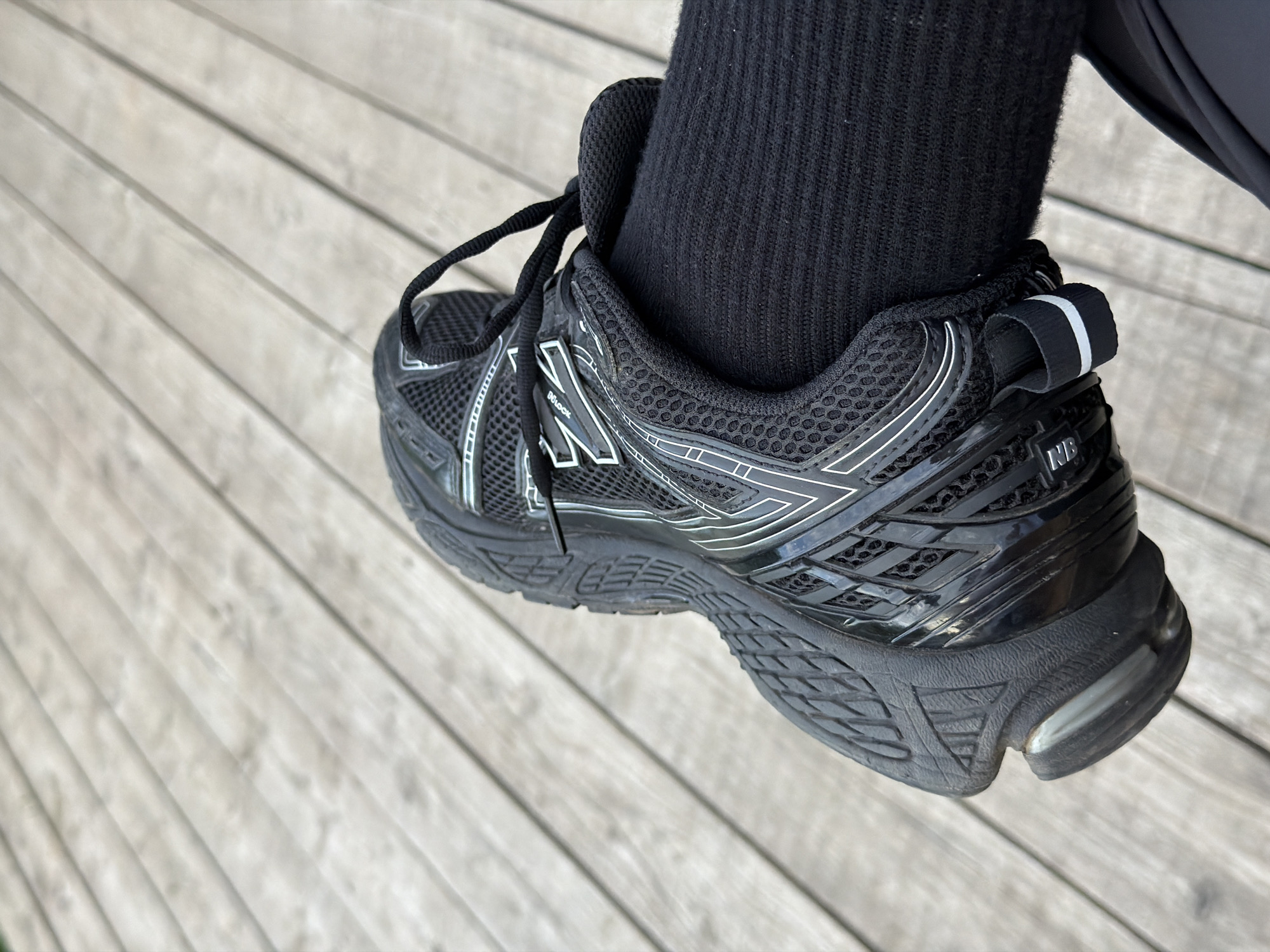 The media file is a close-up, angled shot of a black New Balance athletic shoe and a person's black sock, resting on a light-colored wooden deck. The shoe is detailed with white accents and branding, including "NB" and the New Balance logo on the side. The sole of the shoe shows a textured pattern, and there is a white and black pull tab at the heel. The wooden deck has a weathered, planked appearance and runs diagonally across the frame. The overall lighting suggests it is daytime. The image does not convey a specific activity beyond the shoe being worn and placed on the deck, nor does it provide cues about the weather or time of day beyond daylight. There are no strong emotional indicators present. The location context provided is Bol'shaya Ven'ya, Russia.