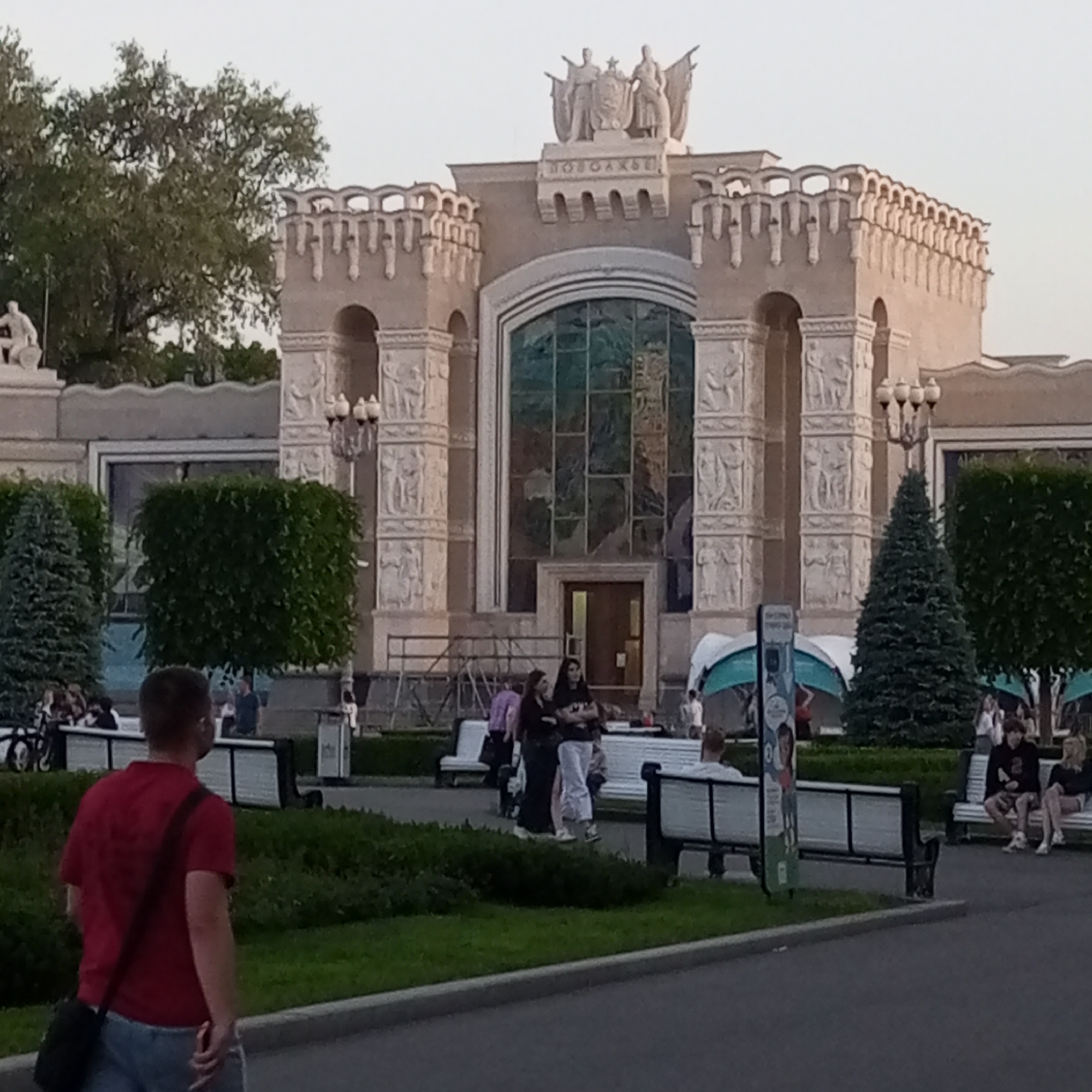 This image depicts Pavilion No. 64 "Optics" (formerly "Kolkhoz Culture" or "Grain") at the VDNKh (Exhibition of Achievements of National Economy) complex in Moscow, Russia. The central subject is the pavilion's ornate facade, constructed of a light brown/beige material.
The building features a large, central arched entrance with a multi-paned, possibly stained-glass, window above a dark entrance door. Two robust pilasters flank the central arch, each adorned with multiple bas-reliefs depicting figures engaged in agricultural activities. Above the central facade, a prominent rooftop sculpture group is visible, consisting of a male figure on the left and a female figure on the right, flanking a central shield emblem with a star and possibly a hammer and sickle, surrounded by banners. Below this sculpture and above the central arch, the Cyrillic word "КОЛХОЗЫ" (Kolkhozes) is inscribed. The roofline above the central section features a decorative parapet with a series of arches. Metal barriers or scaffolding are present directly in front of the building's entrance.
In the foreground, a paved path runs horizontally. On the left, a man with short dark hair, wearing a red short-sleeved shirt and light blue jeans, walks away from the viewer towards the right, carrying a black sling bag over his left shoulder. Green grass and low-lying bushes occupy the space between the path and the pavilion. Several dark green, conical evergreen trees are strategically placed in front of the building.
In the mid-ground, two women stand near the building's entrance; one wears dark clothing, and the other wears a light top and light pants with crossed arms. To the right, a man in a dark hoodie and shorts sits on a white park bench with a woman in a light top and shorts beside him. An ornate, black streetlight with three globes is visible to the right of the central entrance. A tall, slender information sign is positioned on the left, near the man in the red shirt.
In the background, additional green trees, including taller deciduous trees, are visible. Several other indistinct figures populate the park area, some seated on benches or walking. Two light blue and white canopy structures are visible to the right of the pavilion. The overall lighting suggests late afternoon or early evening.