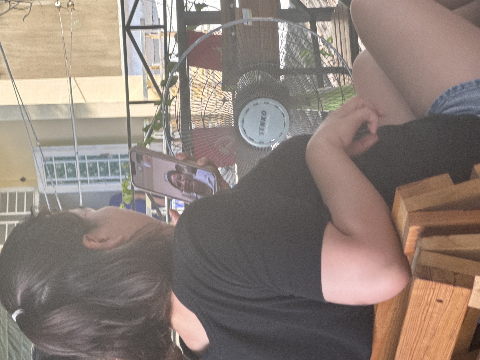 In Bac Nha Trang Ward, Vietnam, a woman with dark hair tied back is seated on a wooden bench. She is holding a smartphone in her right hand, facing away from the camera, and appears to be on a video call with another person whose face is visible on the phone screen. The woman is wearing a black t-shirt and denim shorts. To her right, a large, oscillating fan with "SENKO" written on its center is operating. Behind the woman, a railing and some vegetation are visible. The background suggests an outdoor setting, possibly a balcony or patio, with a building facade and windows.