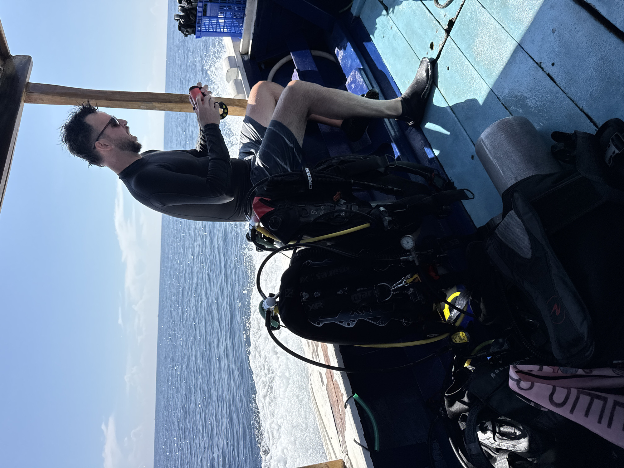 A medium shot presents a male in a wet suit, seated on a blue wooden boat deck with scuba gear. He is facing left with sunglasses on and holding a piece of equipment in his hands, possibly inspecting it. The boat is moving across the water, with the sea and a clear sky visible in the background. The location is Unknown, Indonesia.
