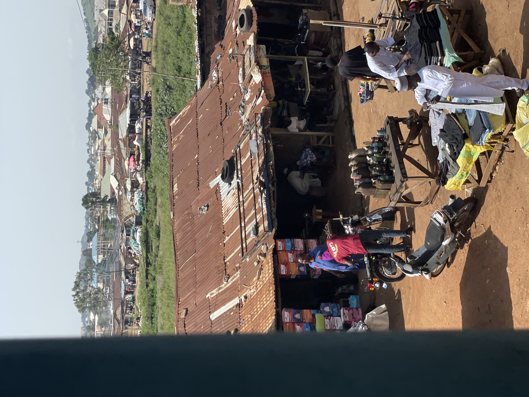 The image depicts an outdoor market scene in a city, likely in a developing country, given the rustic structures and wares. In the foreground, several motorcycles are parked on a dirt ground. A stall displays various items, including what appear to be bottled liquids, possibly fuel or drinks, arranged on a wooden table. Further along, another stall features folded clothing items on tables, with several men engaged in selling or examining the merchandise. One man, dressed in white attire, is meticulously folding a piece of clothing. Behind this stall, a larger structure with a corrugated metal roof, showing signs of rust and wear, serves as a shop or storage area. Several people are visible in and around this structure, with stacks of colorful boxes on display. In the background, more buildings and dwellings extend into the distance, suggesting a densely populated area. The overall environment is characterized by a mix of commercial activity, modest infrastructure, and a clear sky overhead, indicating daytime. The scene has a lively but unpolished feel, typical of a local marketplace.