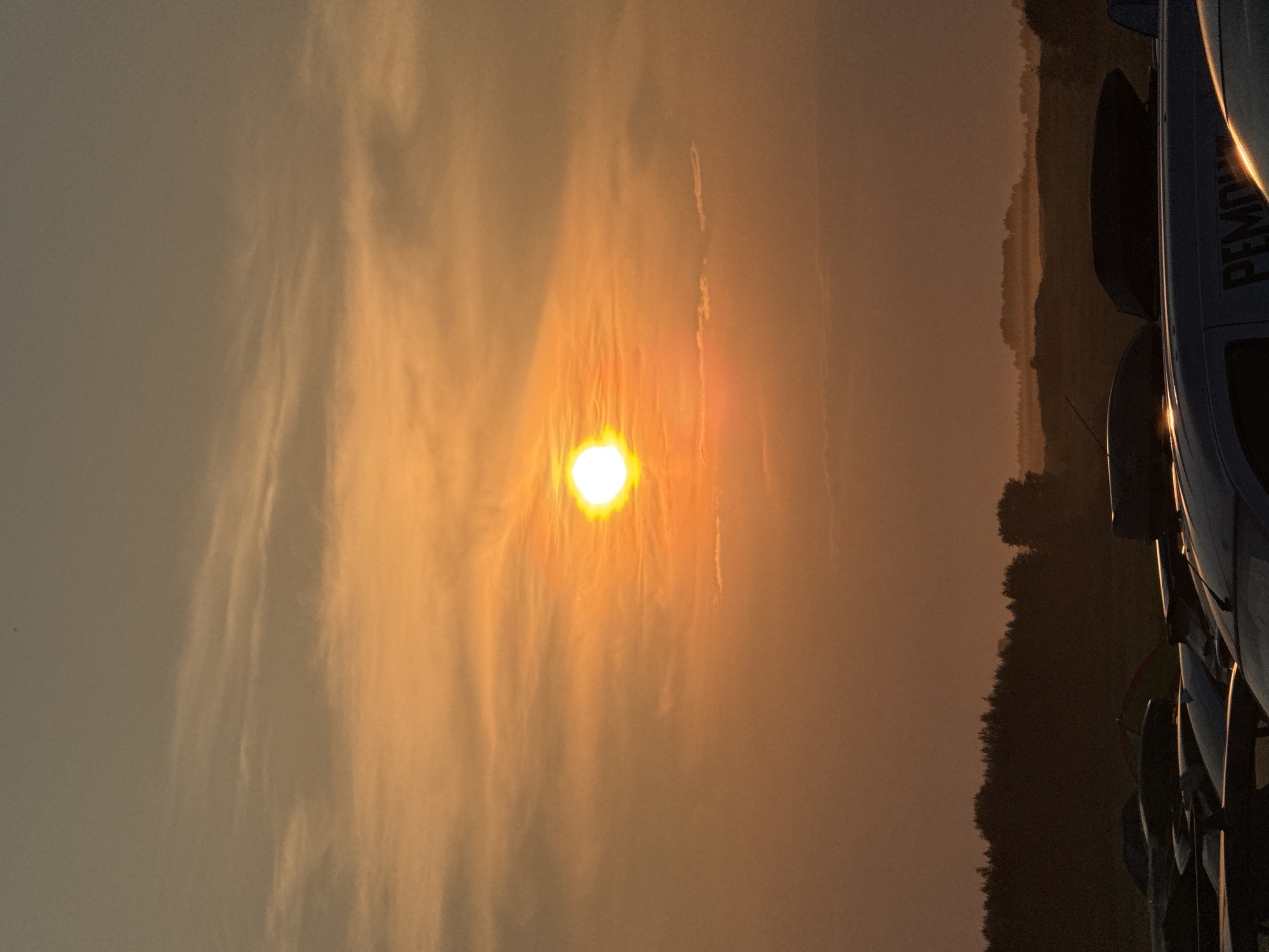 A vertical, eye-level shot captures a sunset over a rural landscape. The sun, a bright, white orb with a hazy orange glow, is positioned in the upper-middle of the frame, surrounded by wispy clouds painted in shades of orange and gray. The sky transitions from a soft orange near the sun to a dusky gray at the top. Below the sky, a dark, silhouetted treeline stretches across the horizon, with some fainter hills visible in the distance. In the foreground, several car roofs, some with roof boxes, are visible, casting long shadows. The overall mood is serene and peaceful, with the warm hues of the sunset creating a tranquil atmosphere.