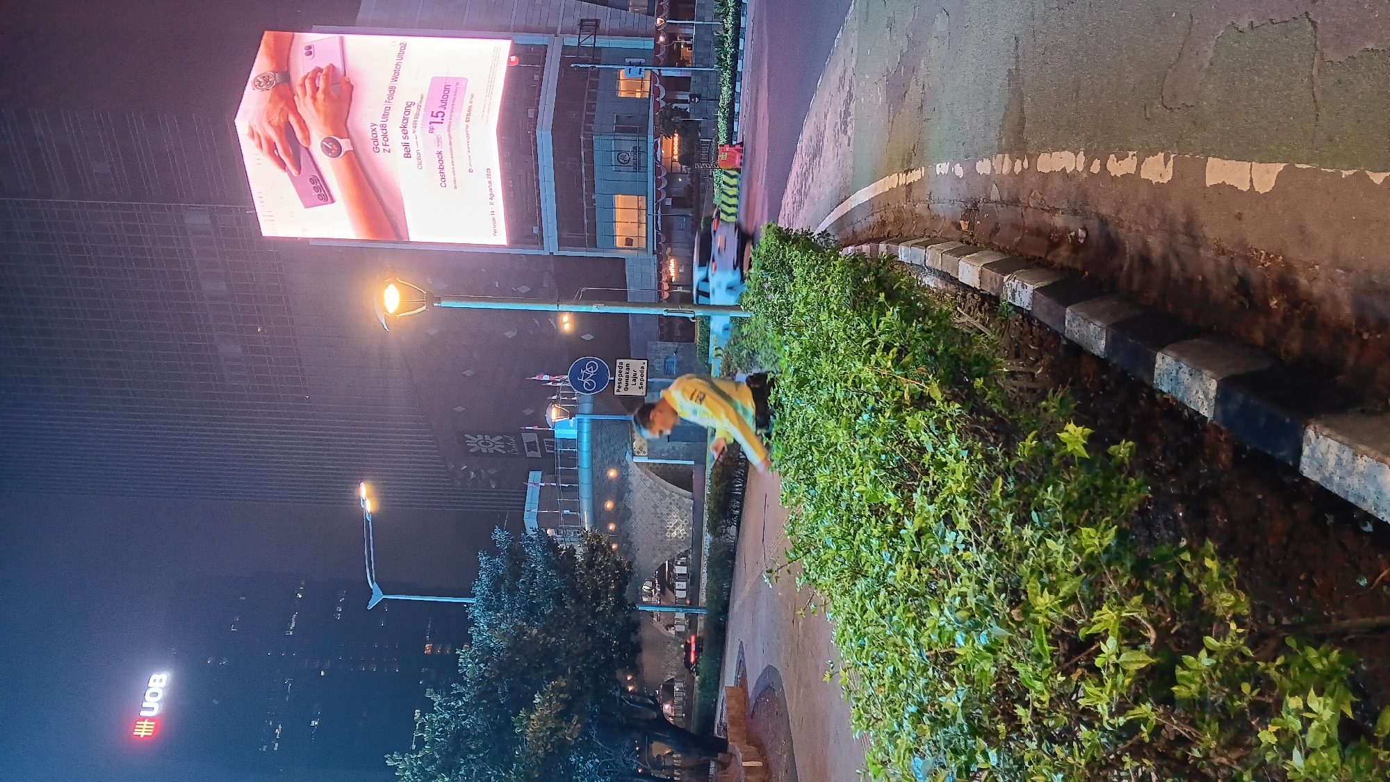 An outdoor, eye-level, medium shot captures a nighttime urban scene in Central Jakarta, Indonesia. A security guard, wearing a yellow shirt and dark pants, stands in the middle ground, looking down at a phone in his hand. He is positioned near a lush green hedge that runs along the lower third of the frame. In the background, a large, illuminated billboard advertises a Samsung Galaxy Z Fold 8 Ultra, featuring images of a watch and a phone. Adjacent to the billboard is a tall building with a UOB logo at the top, indicative of a financial institution. Streetlights and building lights illuminate the scene, casting a warm glow on the wet pavement and creating reflections. A bicycle lane sign is visible on a pole to the right of the security guard, with a sign below it reading "Pesepeda Gunakan Lajur Sepeda" (Cyclists Use Bicycle Lane). A white car is blurred in motion on the right side of the road, suggesting vehicular traffic. The overall atmosphere is that of a busy city at night, with a focus on modern infrastructure and commercial activity.