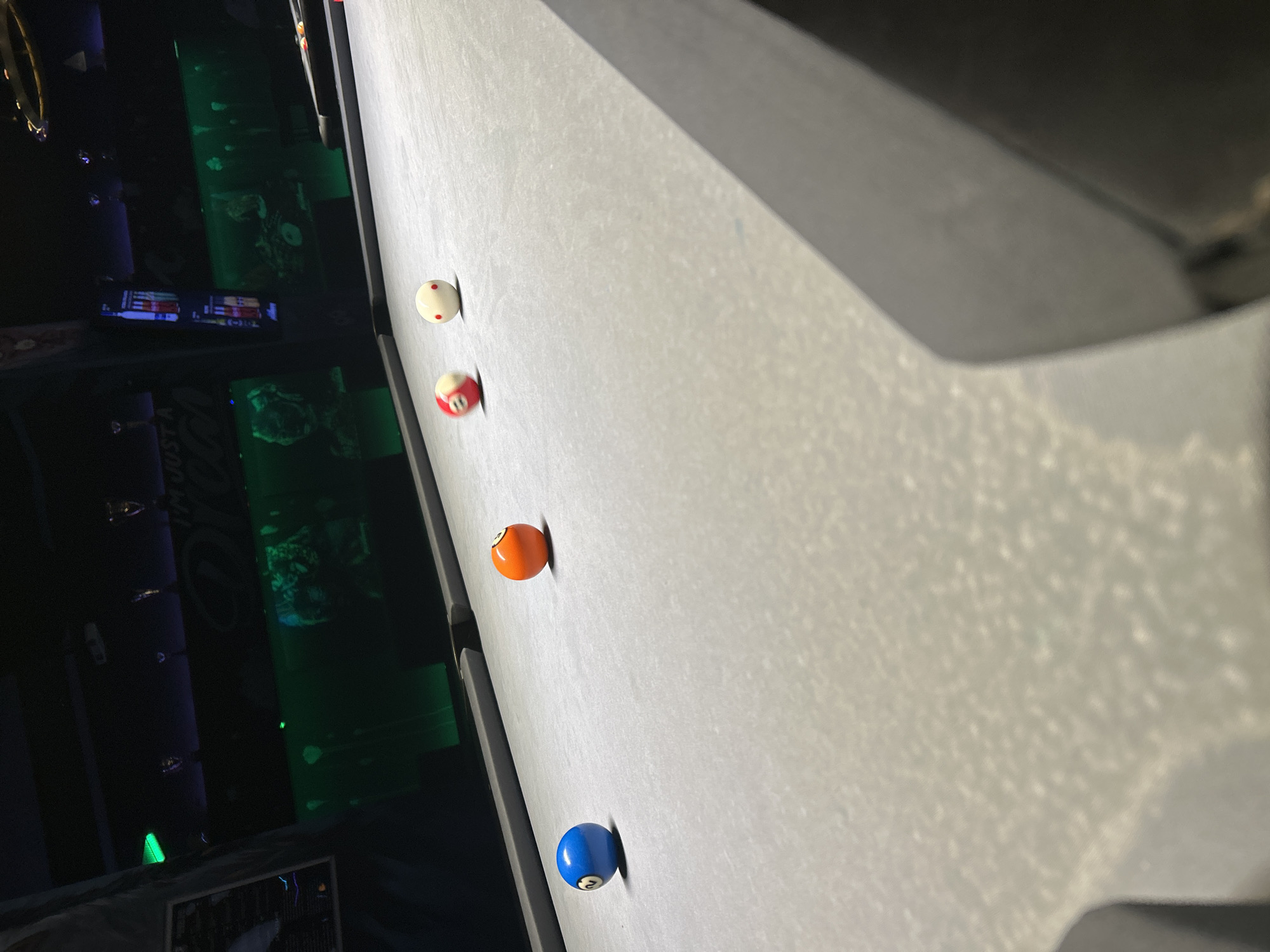 This image depicts a light grey pool table within a dimly lit recreational establishment, specified to be located in Sofia, Bulgaria. Four pool balls are positioned on the table's felt surface: a blue 2-ball, an orange 4-ball, a red-striped ball (appearing motion-blurred), and a white cue ball marked with multiple red dots. The background wall is illuminated by horizontal green light strips, revealing indistinct graphical elements and partial text, notably "I'M JUST A...". Overhead fixtures are faintly visible against the dark ceiling. No individuals are present in the scene.