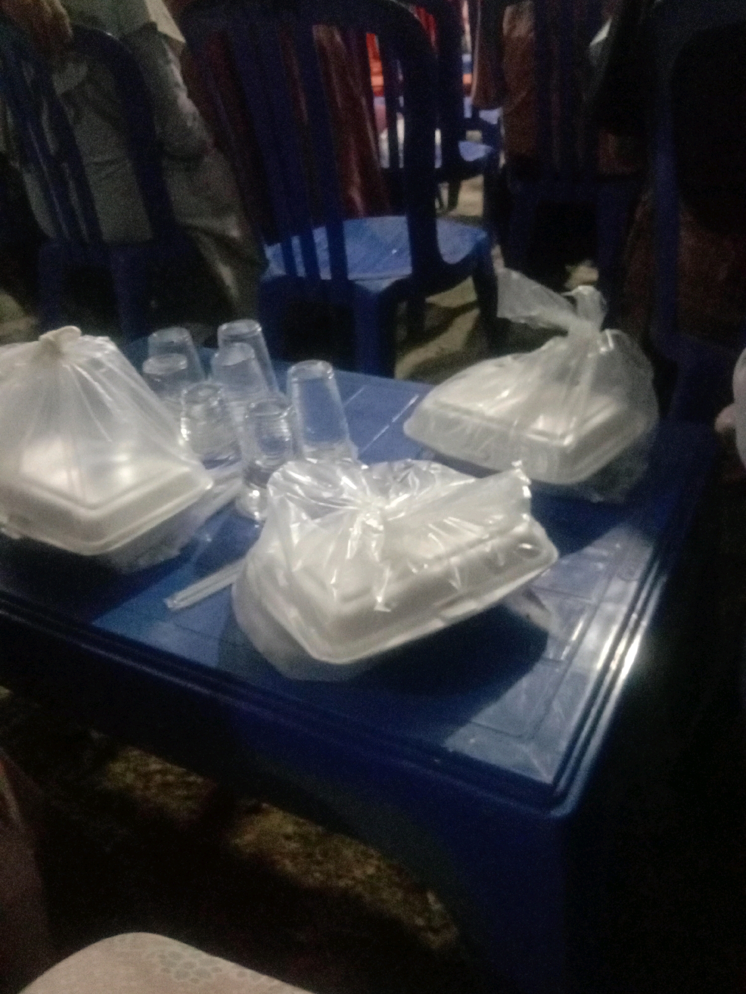 In Grabagan, Indonesia, on a blue plastic table, are two white foam take-out containers, each enclosed in a clear plastic bag. There are also several clear plastic drinking cups and what appear to be straws scattered on the table. The table is set against a backdrop of dark blue chairs and a dimly lit environment, suggesting an outdoor setting, possibly a street food stall or a casual dining area.