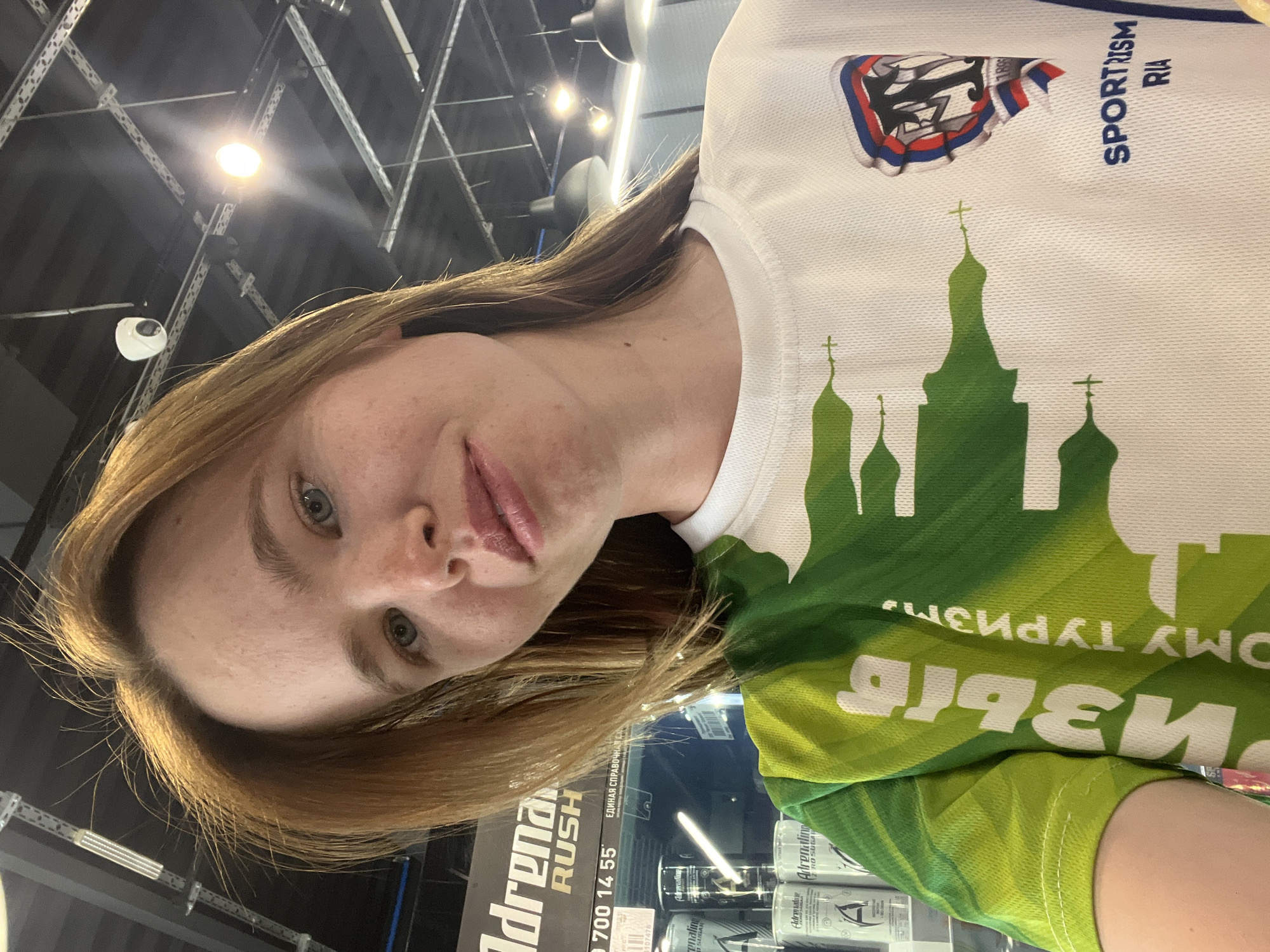 A woman is posing for a selfie. She has light brown hair and blue eyes. She is wearing a white t-shirt with a green silhouette of a Russian Orthodox church. The t-shirt also has a patch with the text "SPORT RISM RIA" and the year 1895. Behind her, there is a shelf with cans of "Adrenaline Rush" energy drinks. Text on the energy drink reads "Adrenaline zero sugar". In the background, there is a metal ceiling structure with overhead lights. The text on the shirt says "ОМУ ТУРИЗМ" which translates to "to tourism" in English. The shirt design suggests she is in Казань (Kazan), Russia, with the depicted churches representing the Kazan Kremlin.