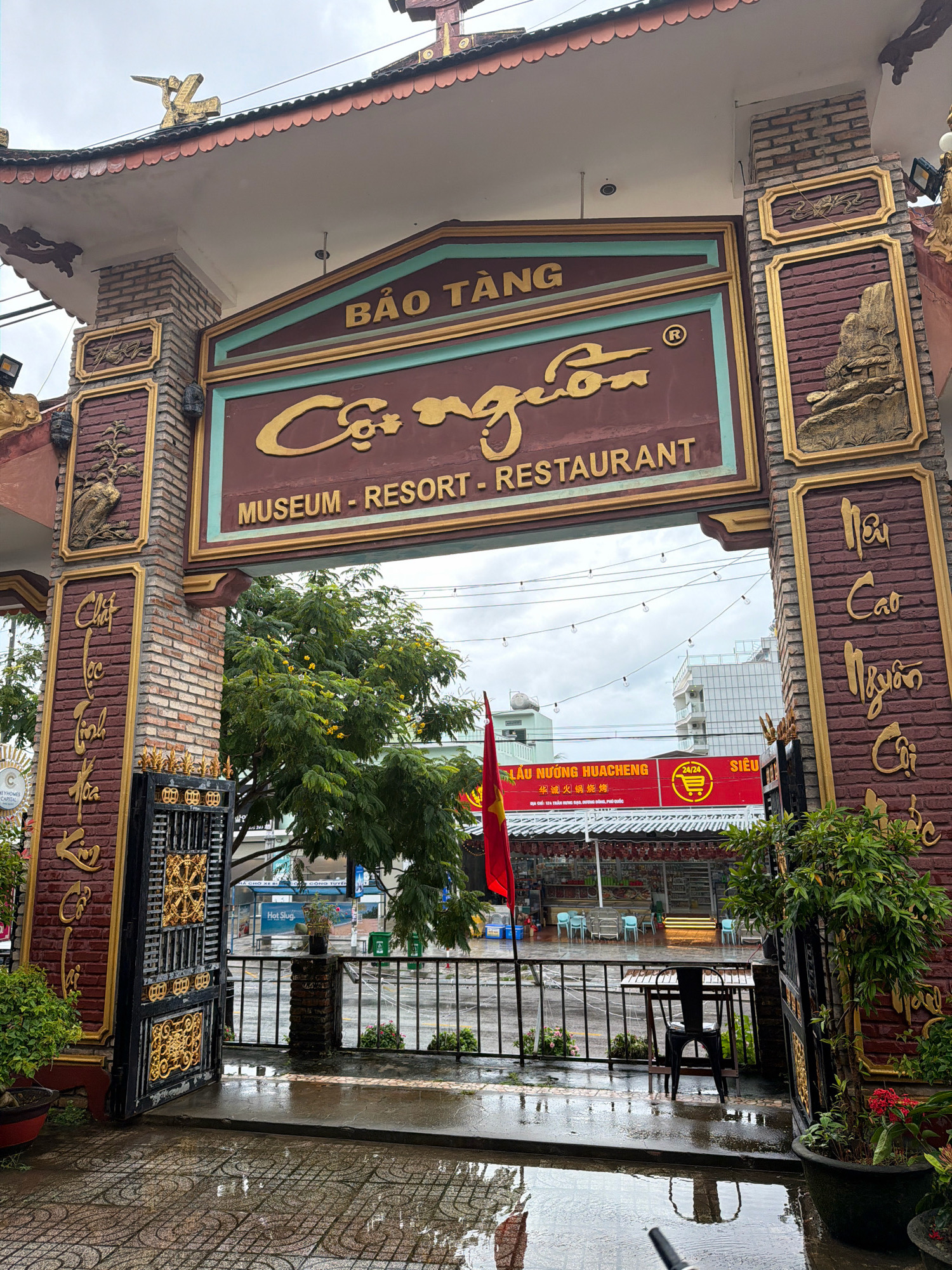 The image shows the entrance to the Báo Tàng Cội Nguồn Museum - Resort - Restaurant in Phú Quốc, Vietnam. The entrance is an ornate archway made of brick and decorated with gold accents. Above the arch, a sign in Vietnamese reads "Bảo Tàng Cội Nguồn" (which translates to "Banyan Tree Origin Museum"). Below this, in English, it says "MUSEUM - RESORT - RESTAURANT". The archway is flanked by brick pillars adorned with gold relief carvings of mountains and traditional Vietnamese calligraphy. A black metal gate with intricate gold designs is open, revealing a pathway leading into the establishment. Beyond the gate, a courtyard area is visible with tables and chairs set up, suggesting an outdoor dining or seating area. A red Vietnamese flag is flying from a flagpole. The ground is wet, indicating that it has recently rained, and puddles reflect the overhead lights and the surrounding environment. Lush green trees with yellow flowers are seen on the right side of the image. In the background, buildings and a cloudy sky can be observed.