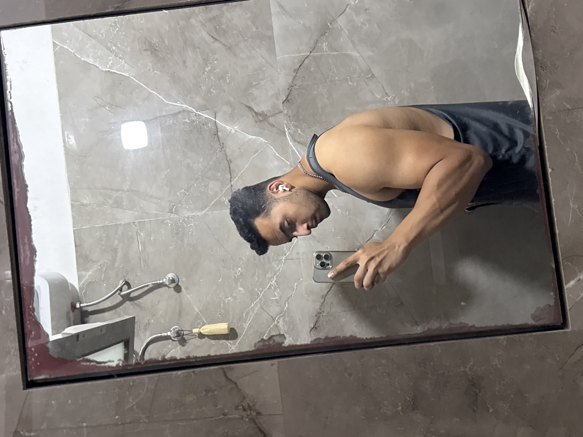 A person is taking a selfie in a bathroom mirror. The bathroom has grey marble tiles. The person is wearing a tank top and has headphones in their ear. They are holding a phone in their right hand. The sink is visible in the mirror. The person appears to be male. The location cannot be determined to be Kolkata, India from the image alone.