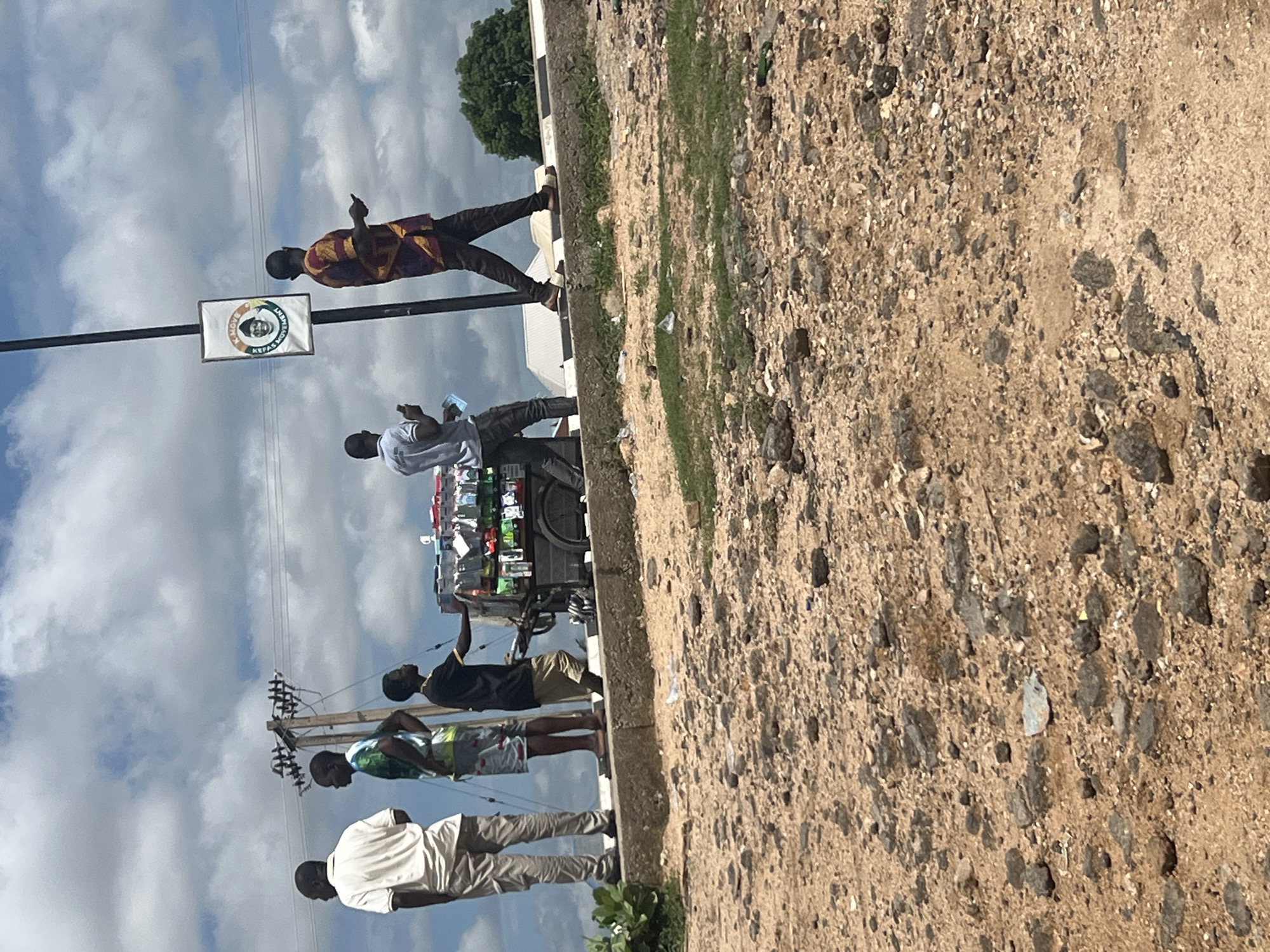 The image captures a daytime roadside scene in Jalingo, Nigeria, under a sky with prominent white cumulus clouds. The foreground consists of dry, reddish-brown earth significantly covered with dark gravel and scattered pieces of plastic litter.
A raised concrete ledge, functioning as a sidewalk or curb, traverses the midground. Standing or walking on this ledge are five males. From left to right:
1. A male, seen from behind, wears a light-colored polo shirt and light-colored cargo pants, standing stationary.
2. Another male, also viewed from behind, wears a multi-colored patterned short-sleeved shirt and light shorts, standing stationary.
3. A younger male, wearing a black t-shirt with a yellow collar and tan shorts, stands with his right hand extended towards items on a two-wheeled vendor's cart.
4. A male, mostly obscured by the cart, wears a light-colored t-shirt and dark pants, appearing to attend to or push the cart. The cart is densely packed with numerous small packaged items, including cell phone cases and other accessories, displayed on external racks.
5. A male, seen from behind, walks towards the right while holding a smartphone in his left hand. He wears a long-sleeved shirt with a distinctive maroon, yellow, and black geometric pattern, paired with dark pants.
A tall, dark utility pole is positioned between the vendor's cart and the walking male. Electrical wires extend horizontally from the pole. A rectangular white banner is attached to the pole, displaying a circular logo with text "K. MOVE" and "LEAD MOVEMENT" encircling a portrait, characteristic of a political campaign. In the background, beyond the concrete ledge, several buildings with light-colored roofs are visible, along with a large green tree on the far right. Green shrubbery grows from the left edge of the concrete ledge. The concrete ledge further to the right exhibits a black and white striped pattern along its edge.