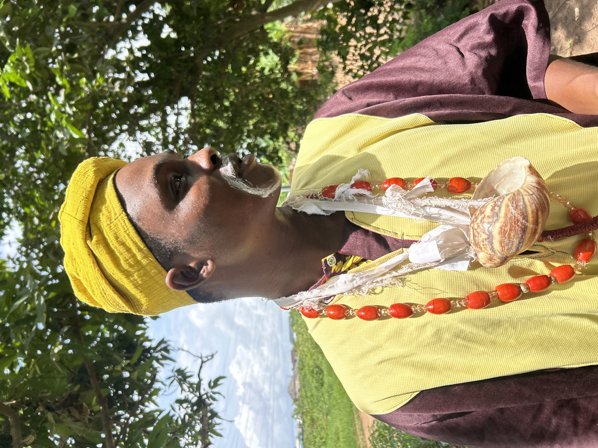 A person wearing a yellow head wrap and a brown and yellow garment stands outdoors. They have white markings on their face, and a necklace of red beads and a large snail shell hangs around their neck. Trees are visible in the background, along with a patch of green grass and a blue sky with white clouds. The location appears to be in a natural setting, not a city.