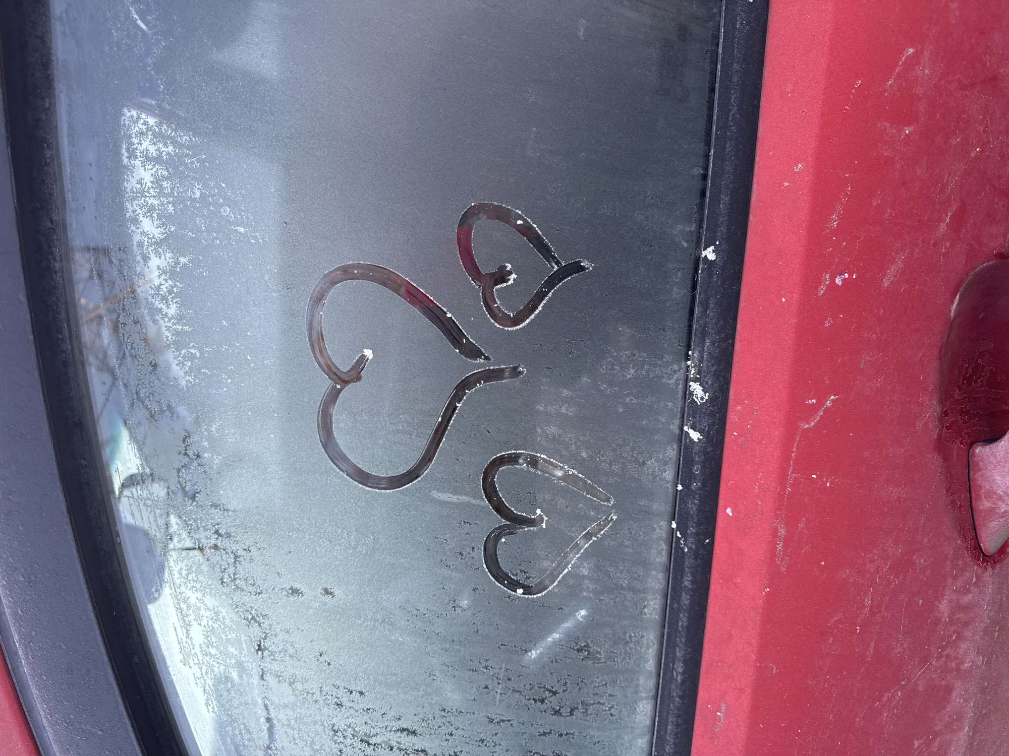 A close-up photograph shows a frosty car window with three heart shapes drawn into the frost. The window is bordered by black trim and part of a red car door is visible to the right. The frost on the window is thin, revealing a gray surface underneath. The location in Chaikovsky, Russia is not evident from the image.