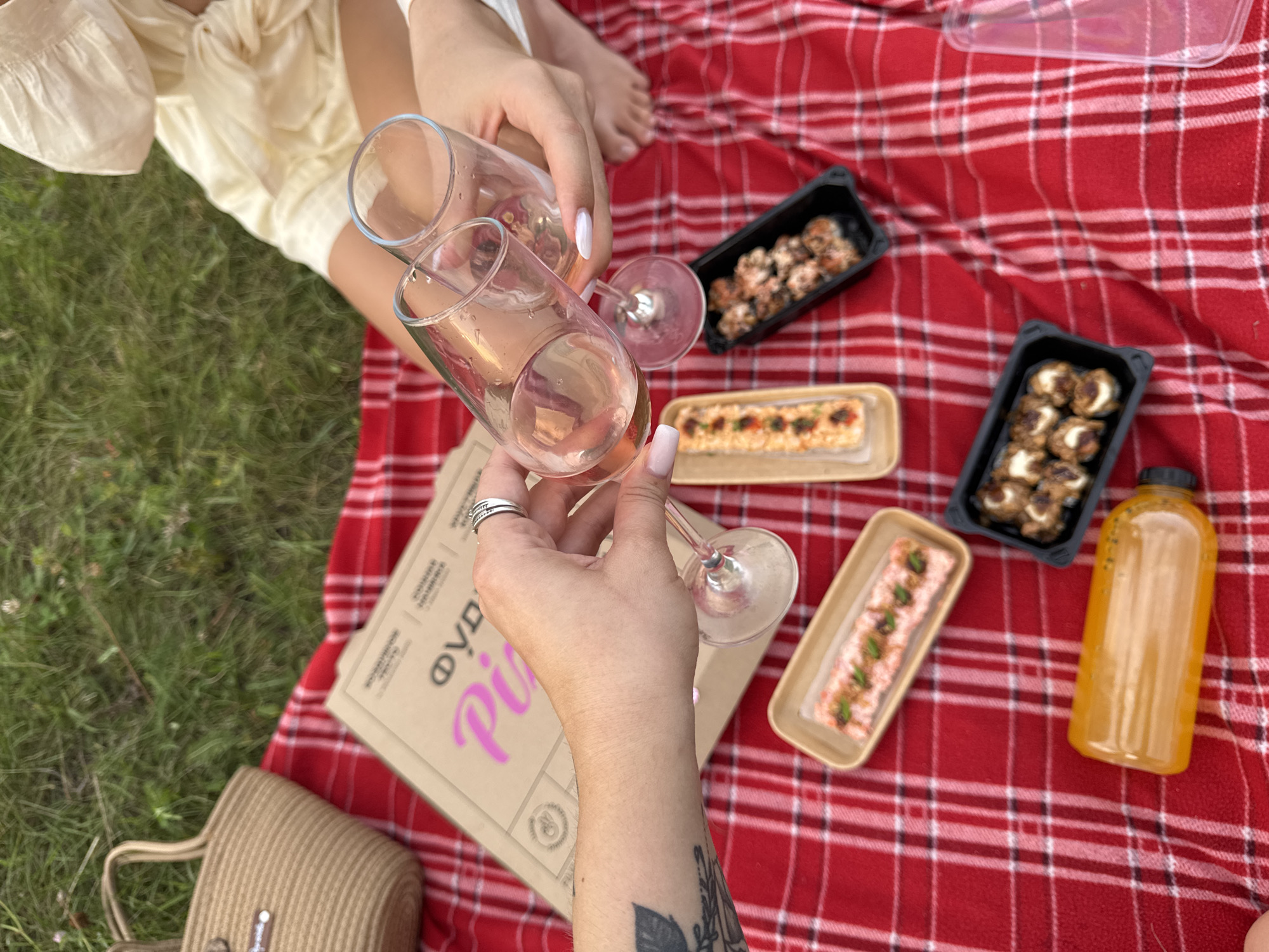 A top-down perspective shot captures a picnic spread on a red and white checkered blanket laid out on lush green grass. Two individuals are present, one's arm extends into the frame from the left, holding a wine glass filled with pink liquid, their hand adorned with rings. Another person's hands are seen on the right, also holding two wine glasses, one partially filled with the same pink beverage, and a glimpse of their cream-colored puffed-sleeve top and bare feet is visible.
The picnic arrangement features a cardboard pizza box with pink lettering that appears to read "Pizza," a beige woven tote bag, and several food containers. These containers hold various food items, including what looks like sushi or sashimi in elongated rectangular dishes, and small, bite-sized rolls or appetizers in black rectangular containers. A bright orange beverage bottle lies on its side in the foreground. The setting is outdoors, likely in a park or grassy area, suggesting a casual gathering. The image does not contain any specific markers or context to identify the location as Tolyatti, Russia.