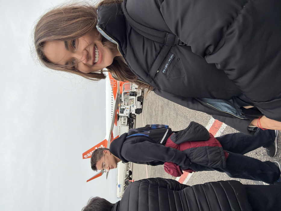 The image depicts an outdoor scene on an airport tarmac at Lyon-Saint Exupéry Airport in Colombier-Saugnieu, France, under an overcast sky. In the foreground on the right, a young woman with long brown hair smiles, looking towards her right, off-camera. She wears a black puffer jacket featuring a "PRADA - MILANO" triangular logo patch on the left chest and a red string bracelet on her right wrist. Behind her, in the mid-ground, a young man with short brown hair and eyeglasses wears a dark hooded sweatshirt and dark trousers, carrying a dark backpack and a red and black puffer jacket over his left arm. He is looking to his left. Partially visible on the far left foreground is the back of a third individual wearing a dark puffer jacket. In the background, an EasyJet aircraft, white with an orange tail and "easyJet" logo, is parked on the tarmac. Various white ground support vehicles, including a fuel tanker truck with "World Fuel Services" visible, are also present on the tarmac. Red and white markings are visible on the tarmac surface. The individuals appear to be walking in an open-air airport environment.