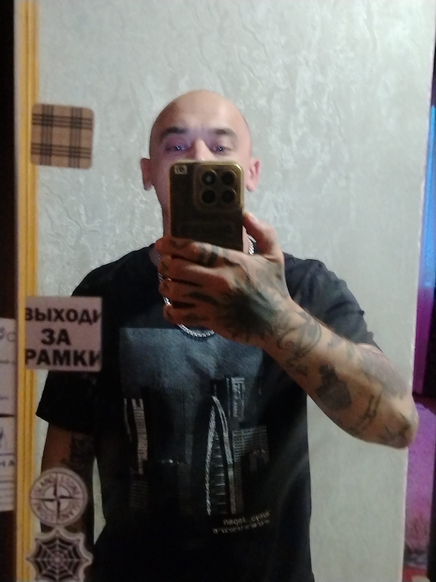 A man with a shaved head and visible tattoos on his arms takes a mirror selfie indoors. He is wearing a dark t-shirt with a graphic print. In the background, a textured wall is visible, along with a plaid sticker and a sticker with the Russian text "ВЫХОДИ ЗА РАМКИ," which translates to "go beyond the frame." Another circular sticker with a compass rose design and a spiderweb design are also on the surface where the photograph is being taken. The man is holding a smartphone in his right hand, angled to capture his reflection.