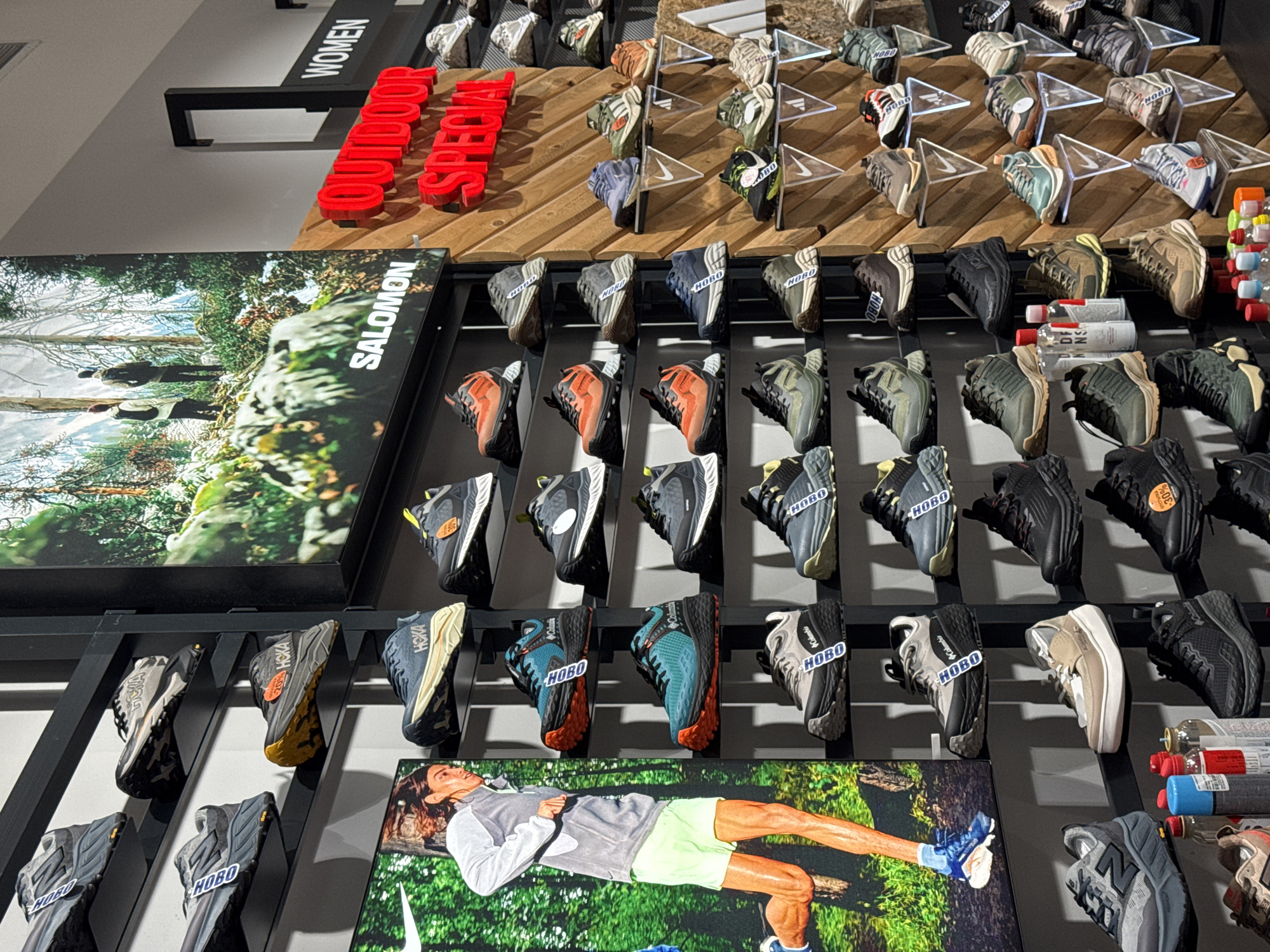 This image captures an indoor retail display of outdoor and athletic footwear, likely in a sports or outdoor equipment store in Sofia, Bulgaria. The primary subjects are numerous pairs of trail running and hiking shoes, arranged meticulously on multi-tiered black metal racks and wooden shelves.
The shoes are varied in brand and color, with prominent brands including Hoka, Columbia, and New Balance (indicated by "NB" on some shoes). Many shoes have small tags, some of which are misidentified as "HOBO" by OCR but appear to be labels or small brand details. Price tags are visible on some items, with one clearly showing "-30%". Alongside the shoes, some outdoor accessories like water bottles and small gas canisters are displayed.
Large promotional display boards are integrated into the setup. One vertical board on the left features a scenic image of two people hiking through a lush forest, with the brand "SALOMON" visible. Another horizontal board on the lower right shows a man lying on a bed of green foliage, wearing athletic attire and shoes, with a partial Nike swoosh logo visible at the bottom.
Prominent red dimensional letters spell out "OUTDOOR SPECIAL" on a wooden shelf, emphasizing the category of products. Above this, a black sign with white letters reads "WOMEN", indicating a section of the display. The overall setting is brightly lit, creating a clear view of the merchandise.