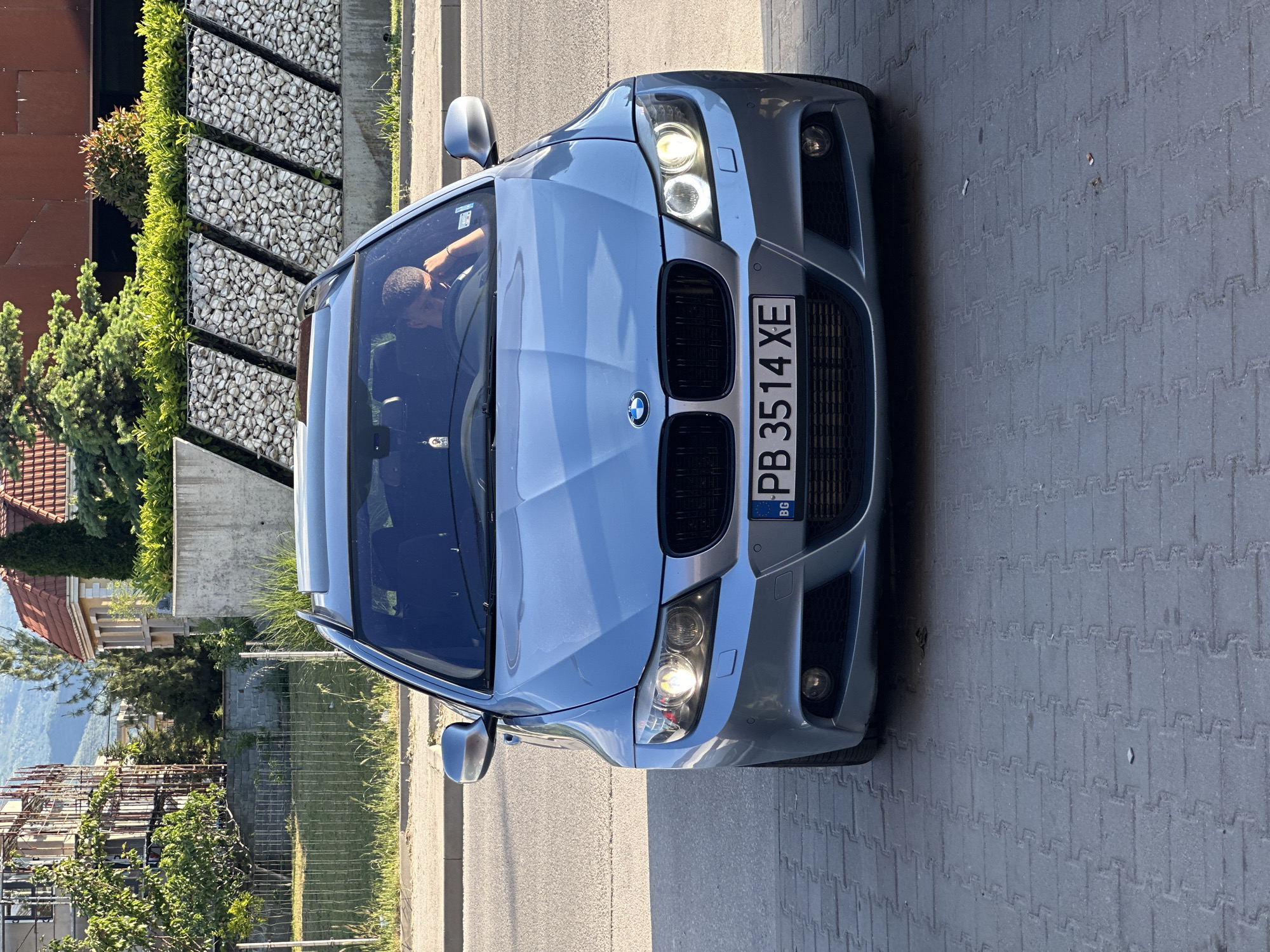The image shows a silver BMW sedan parked on a gray brick road. The car has a Bulgarian license plate "PB 3514 XE." A person is visible inside the car. In the background, there are buildings, green shrubbery, and a rock wall. The scene appears to be in a residential area. Given the Bulgarian license plate, it can be assumed the location is Plovdiv, Bulgaria.