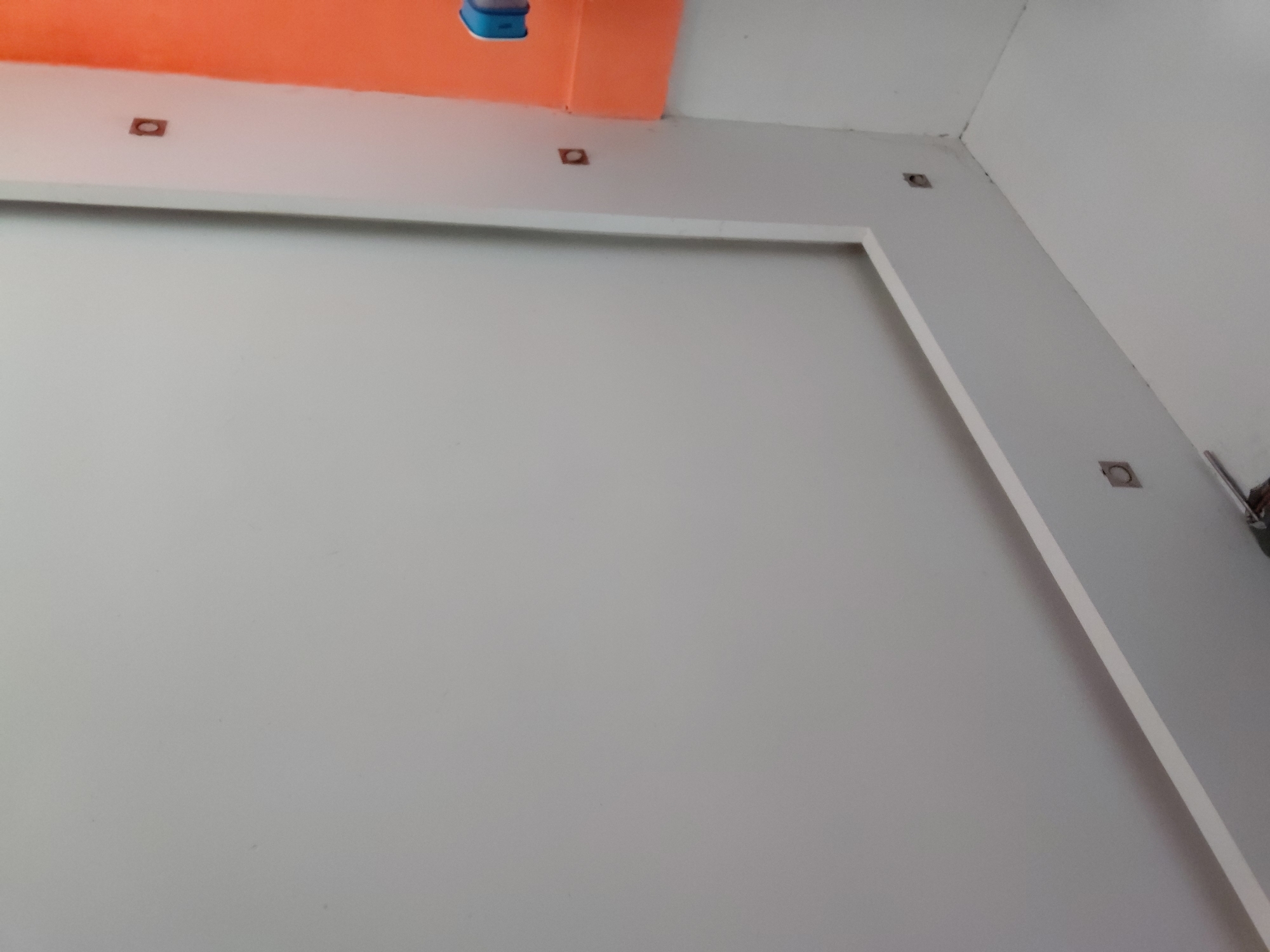 This image captures an interior architectural detail, likely within a room in Santa Ana, Venezuela, presenting a view looking upwards into a corner. The primary subjects are the walls and ceiling.
The ceiling features a striking section painted in a vibrant orange, contrasting with a lower white soffit or beam that runs along its edge. On this white soffit, several small, square-shaped cutouts are visible, likely intended for recessed lighting fixtures. One such cutout is clearly seen towards the left, appearing empty or with a dark inner casing. A similar cutout is visible further to the right under the orange ceiling. A partial view of a blue, cylindrical or box-shaped object, possibly a light fixture or a decorative element, can be seen hanging from the orange ceiling section near the top right.
The main wall in the foreground is a plain light gray or off-white, featuring a large, recessed rectangular panel or frame, creating a layered effect. The top edge of this panel runs horizontally across the image. The side wall to the right is also light gray and extends into the corner. Along this right-hand wall, another series of square cutouts for recessed lights is visible, with at least two clearly identifiable. Near the upper right corner of the image, where the walls and ceiling meet, a metallic pipe or conduit runs vertically along the wall, adjacent to one of the recessed light cutouts.
No people or activity are depicted. The scene is static, focusing on the structural and decorative elements of the room. The lighting appears to be ambient, as the recessed lights themselves do not seem to be installed or active. There is no visible text in the image.