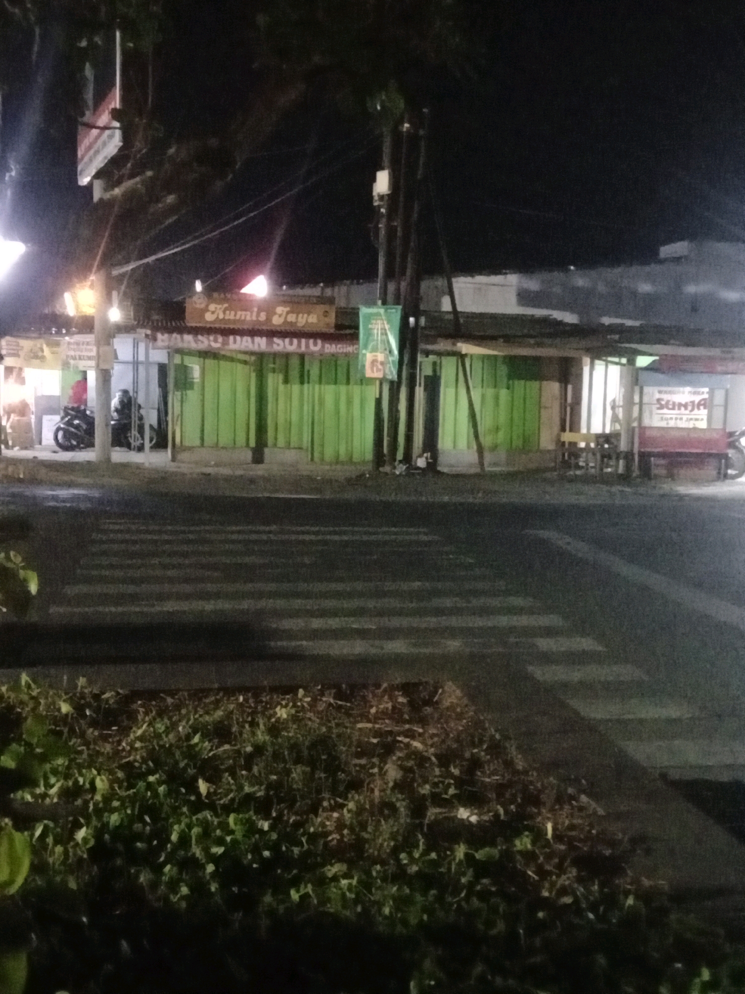 The image depicts a street scene at night, likely in Banyumas, Indonesia, focusing on a row of small shops or food stalls. The prominent signage reads "Kumil Jaya" and "Bakso dan Soto Daging," indicating a food establishment. To the right, another stall displays the name "SUNJA." Several motorcycles are parked in front of the stalls. A pedestrian crossing is visible in the foreground, with a patch of vegetation to the lower left. The lighting suggests it is nighttime, with artificial lights illuminating the area. The overall impression is of a typical roadside commercial area in a town or city.
