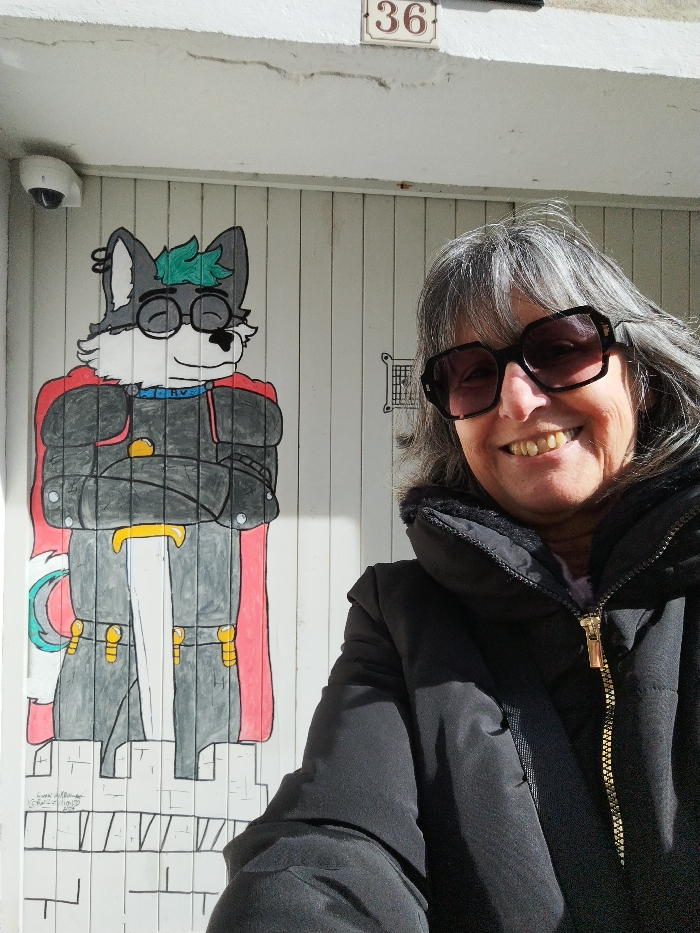 A woman wearing sunglasses and a dark jacket stands in front of a white door. On the door, a cartoon drawing of a gray wolf-like figure dressed in a knight's armor with a red cape is visible. The figure has glasses and a small green tuft of hair. The number 36 is displayed above the door. There is also a security camera mounted near the top corner.