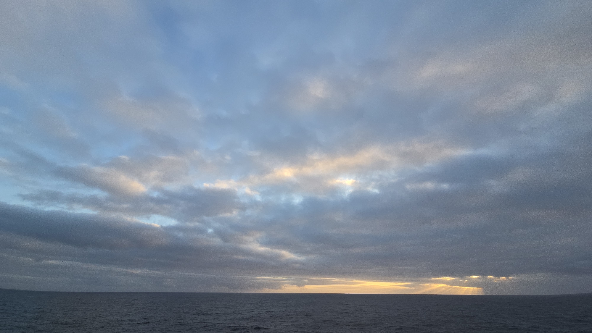 The image depicts a seascape under a cloudy sky. The ocean is relatively calm, with subtle ripples on the surface. The sky is predominantly covered with layers of clouds, varying in color from gray to blue, with some areas illuminated by sunlight. The horizon line is visible, separating the ocean from the sky. Rays of sunlight are visible in the right side of the image. The location is Unknown, Unknown.