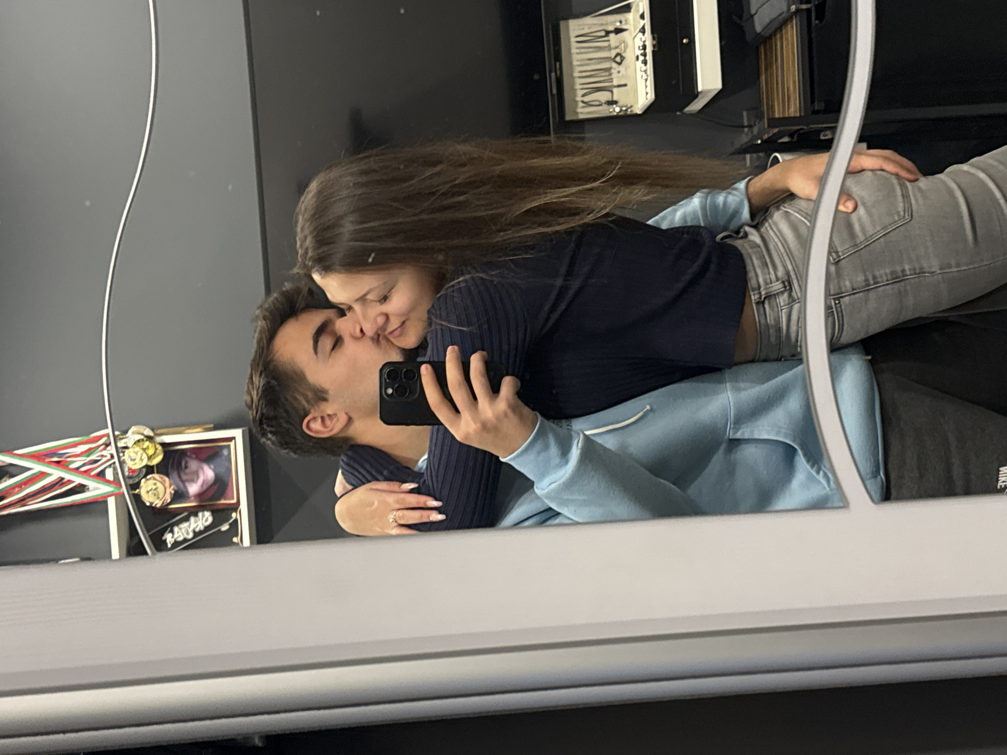 In a mirrored reflection, a young couple is embracing. The man, with short dark hair, is kissing the woman on the nose while holding a smartphone up to capture the moment. The woman, with long brown hair, is smiling. She is wearing a dark blue ribbed shirt and light gray jeans. The man is wearing a light blue hoodie and dark pants. The setting appears to be indoors, possibly a residential or commercial space. Behind them, visible in the reflection, are shelves or storage units. There is a framed photo with medals and ribbons visible on the shelf.
Location cannot be confirmed.