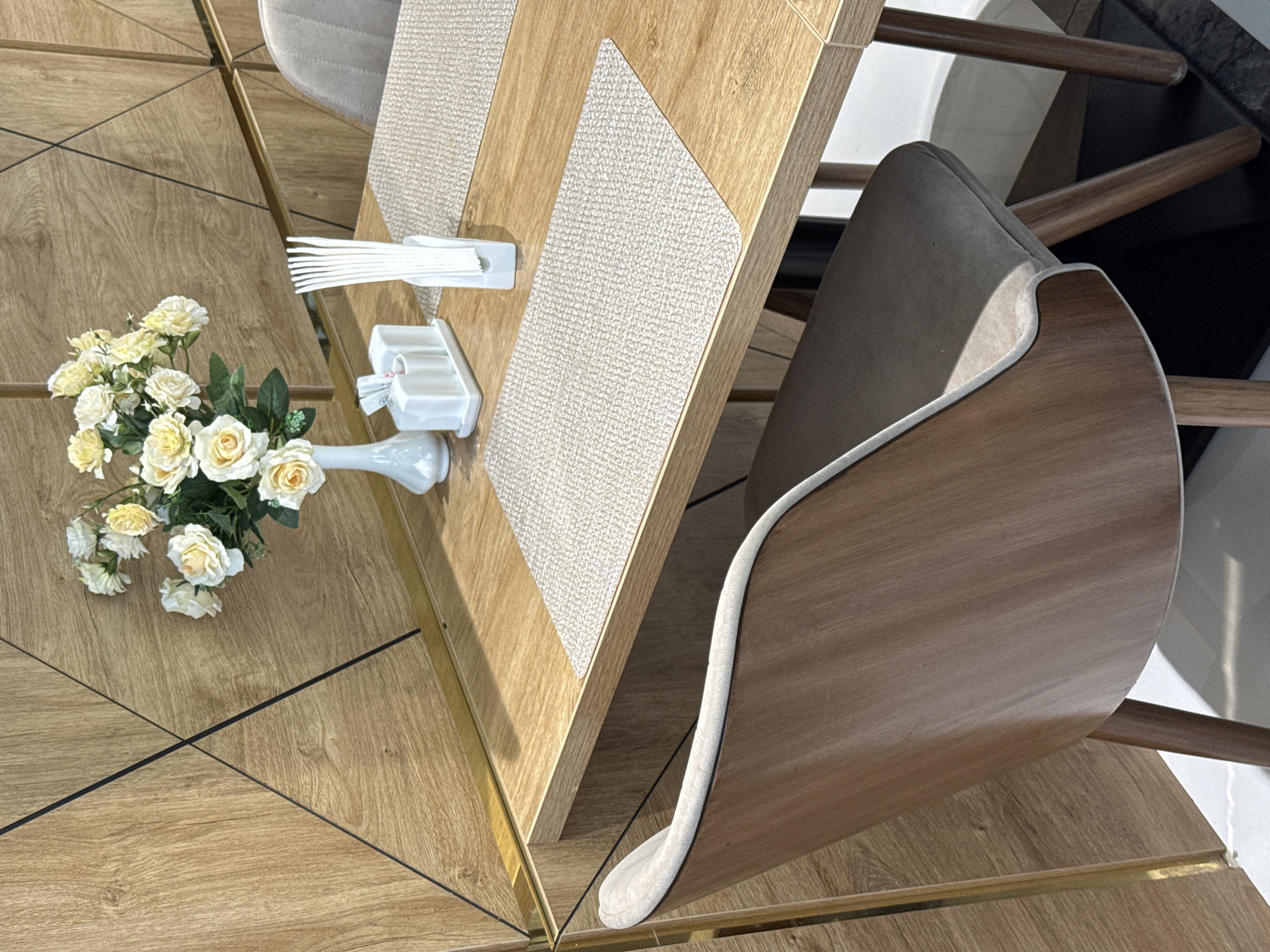 The image captures a high-angle view of a dining table setting, featuring a floral centerpiece, cutlery, and chairs. The table has a wood-grain finish with a gold-toned border, and patterned placemats are positioned on its surface. The centerpiece consists of a bouquet of white and yellow roses in a white vase. A set of white cutlery rests on the table next to a dispenser. A chair with a curved wooden back and a light-colored seat is visible, partially obscuring the table. The flooring has a wood-grain pattern with dark lines forming geometric shapes. The background includes hints of white surfaces. The location cannot be determined based on this image alone, there is no indication it is in Tashkent, Uzbekistan.