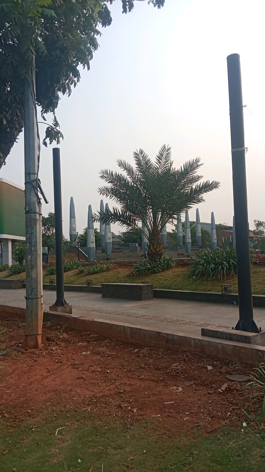 The image depicts an outdoor scene in the city of Bekasi, Indonesia, likely at a public park or monument. In the foreground, there is a paved pathway with a concrete border, adjacent to a patch of reddish-brown soil and green grass. Several tall, dark cylindrical poles are positioned along the pathway, two of which are prominently visible on the left and right sides of the frame.
In the middle ground, a cluster of tall, slender, grey monuments with rounded tops are arranged in a semi-circle. These monuments appear to be decorative or commemorative, with some featuring images or inscriptions on their surfaces. A date palm tree stands in front of these monuments, its fronds fanned out. Lush green bushes and manicured grass surround the monuments and the palm tree, creating a landscaped area.
In the background, a light-colored building with green accents is partially visible on the left, suggesting it might be a visitor center or part of a complex. The sky is overcast and bright, indicating it might be daytime. The overall scene suggests a well-maintained public space, possibly with a historical or cultural significance. No people are visible in the image.