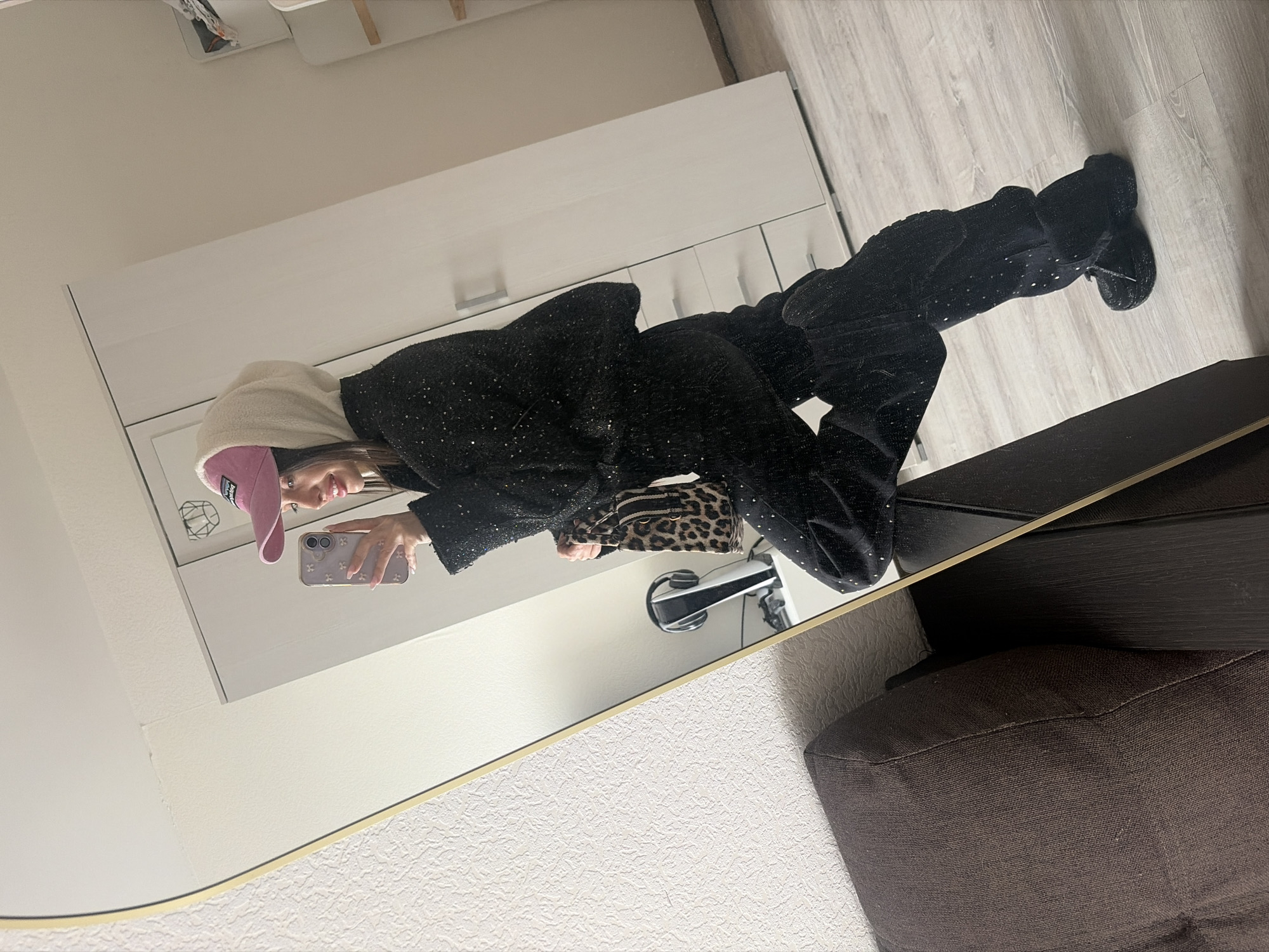 A young woman is taking a selfie in a mirror. She is wearing a pink cap, a beige hood, a black, sparkly jacket, black pants, and black shoes. She is holding a phone with a patterned case. She also has a patterned purse on her shoulder. Behind her is a white dresser, and to the side is a dark-colored sofa. The picture appears to be taken indoors. There is no information to suggest this is located in Saint Petersburg, Russia.