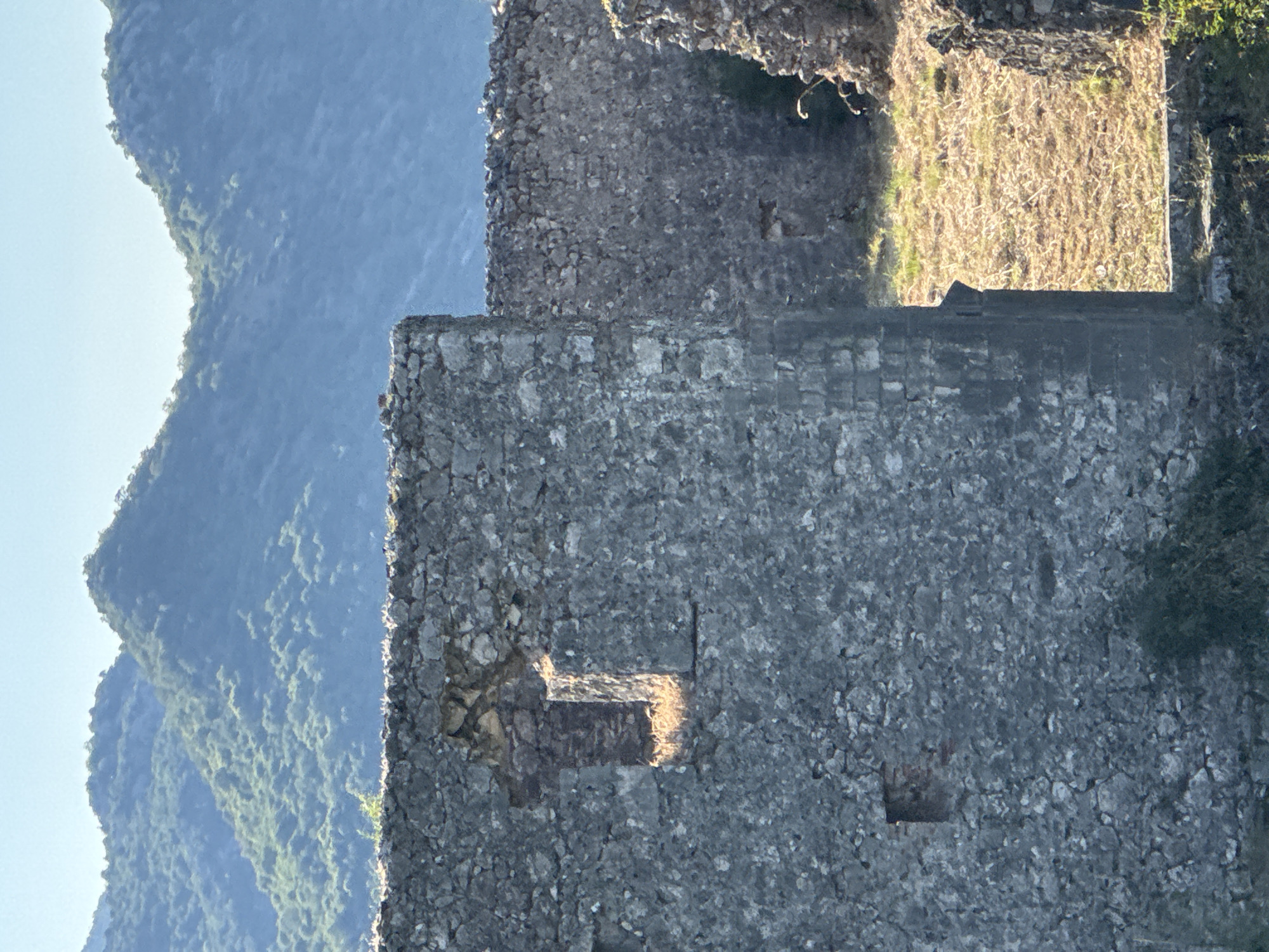 The image depicts the ancient stone walls of the Vrmac fortress in Boljevići, Montenegro. The weathered gray stones form substantial structures, with one section of wall prominently featuring a narrow, cross-shaped opening, likely a loophole for observation or defense. Sunlight illuminates the interior of this opening and casts a warm glow on the dry, golden grass and earth visible beyond another opening in the wall. In the background, lush green mountains rise, their slopes covered in dense vegetation, under a clear, pale blue sky. The scene conveys a sense of historical depth and natural beauty.