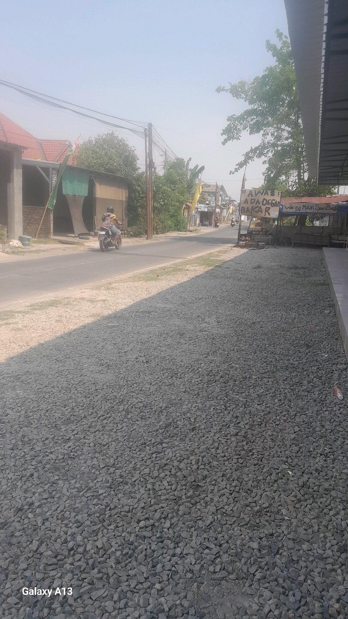 This image depicts a street scene in Kirig, Indonesia, characterized by a dusty, light-colored sky and dry conditions. A paved road runs horizontally across the frame. On the left side of the road, there are buildings with red-tiled roofs and brick structures. A person is seen riding a motorcycle in the middle of the road, heading away from the viewer. To the right of the road, there is a warung, or roadside stall, with a sign that appears to say "AWAS ADA DEGAN BAKAR" which translates to "Beware of Grilled Banana." Adjacent to the warung, a sign for "Warung Makan Sayo Bae" is visible. The foreground of the image is dominated by a large area covered in grey gravel. The overall impression is of a typical, sun-drenched roadside in a rural or semi-rural Indonesian setting.