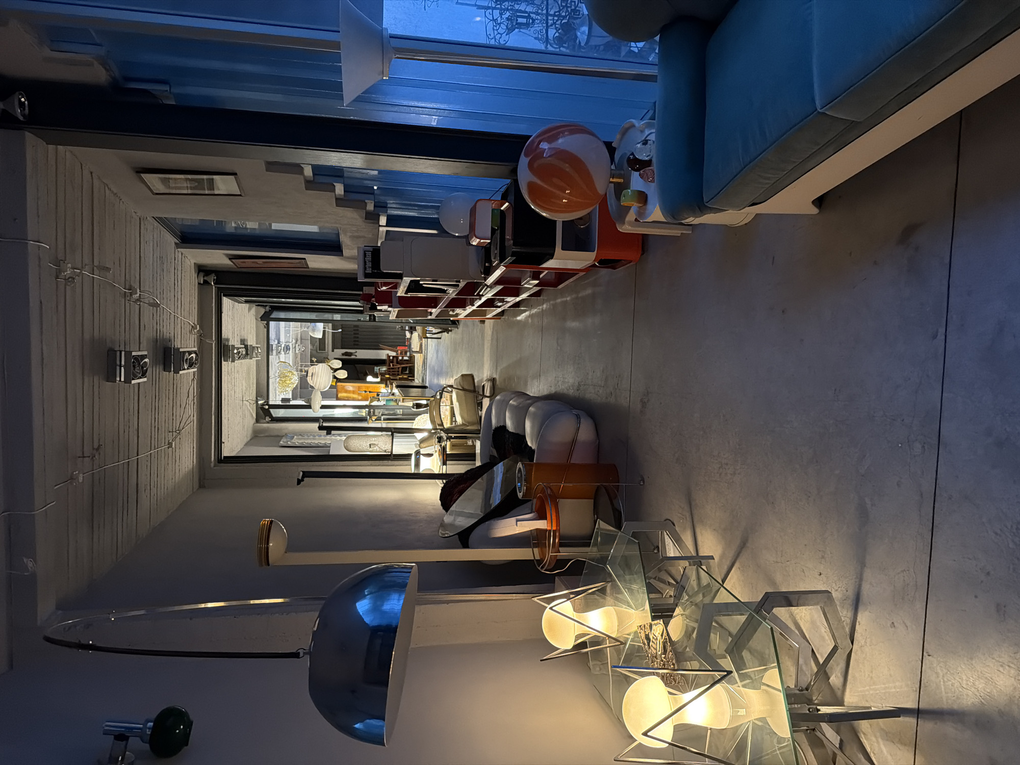 The image depicts an interior space featuring a collection of design objects and furniture. The location is possibly in Buenos Aires, Argentina, based on stylistic cues. The walls are a neutral, textured material, and the floor is a smooth, gray surface. Light fixtures of various designs are prominent, including a large, chrome dome lamp, a multi-bulb geometric lamp, and other standing lamps. The furnishings consist of a blue couch, a red and white shelving unit with various decorative items, and assorted chairs and tables. The perspective suggests a retail or showroom setting where design objects are displayed. No people are visible. The lighting is a mix of artificial and natural, creating a bright and airy ambiance.