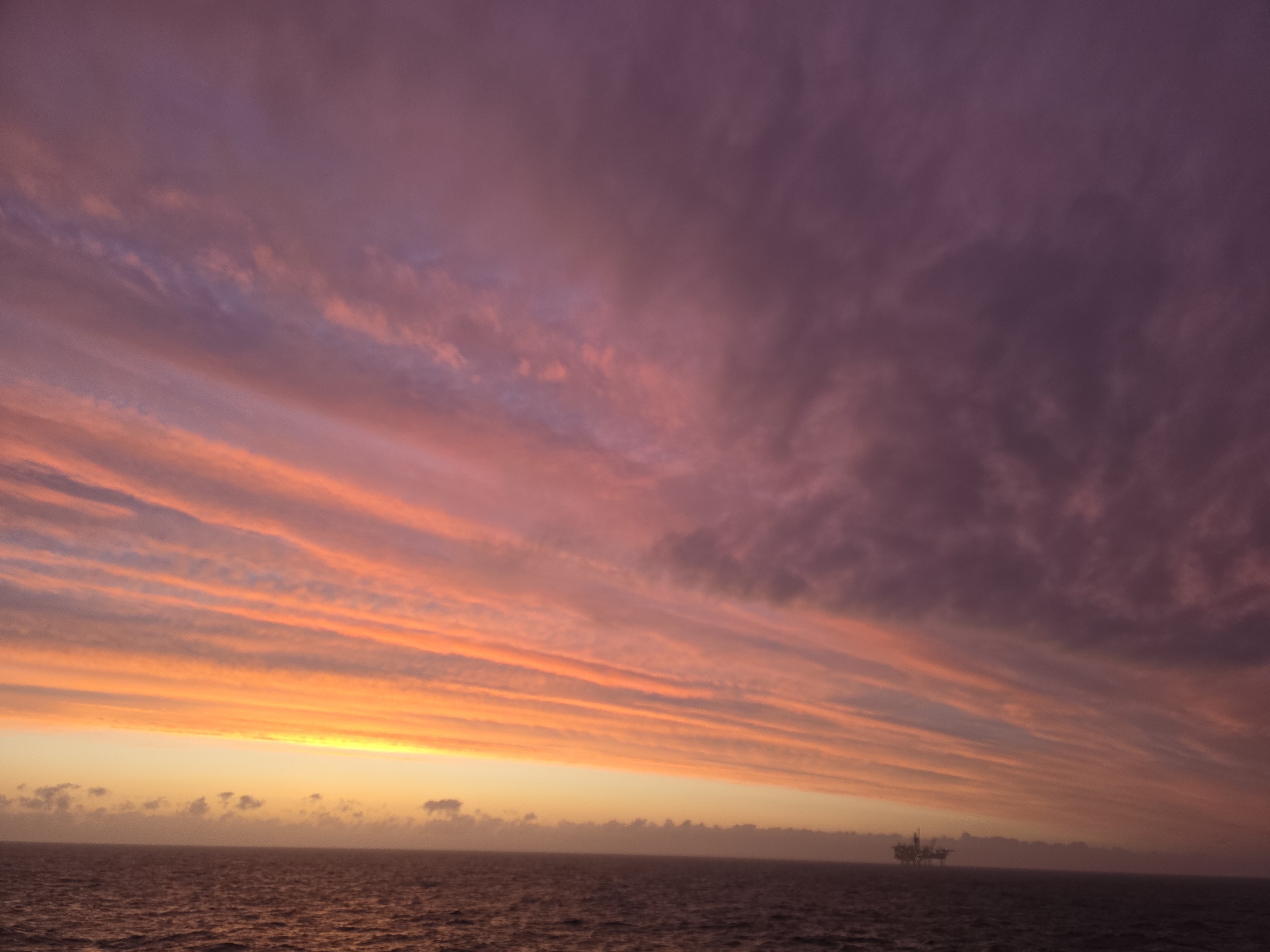 The image depicts a sunset over the ocean. The sky is filled with colorful clouds, ranging from orange and yellow near the horizon to pink and purple above. The clouds are streaky and layered. The ocean is dark with slight ripples on the surface. In the distance, there is what appears to be an oil rig or platform. The location is unknown.