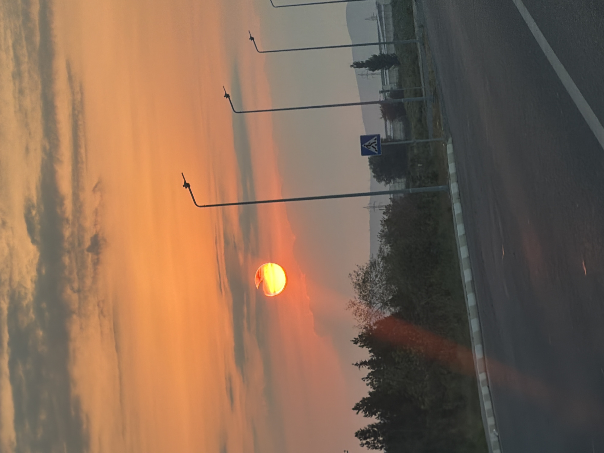 An outdoor, eye-level, medium shot captures a vibrant sunset over Qaragüney, Azerbaijan. The sky is painted with hues of orange and yellow, with scattered gray clouds obscuring parts of the sun, which is partially visible as a bright orange disk with a glowing halo. In the foreground, a paved road with a white line marking the lane curves to the right. A curb with alternating white and gray segments runs along the edge of the road. Beyond the curb, a line of dark green trees forms a dense forest, with some lighter green foliage interspersed. Several tall, slender streetlights with single fixtures stand in a row along the edge of the road. A blue and white pedestrian crossing sign is visible between two of the streetlights. In the distance, faint outlines of hills or mountains can be seen, along with power lines and some industrial-looking structures. The overall scene depicts a quiet, possibly rural or semi-urban, setting at dusk.