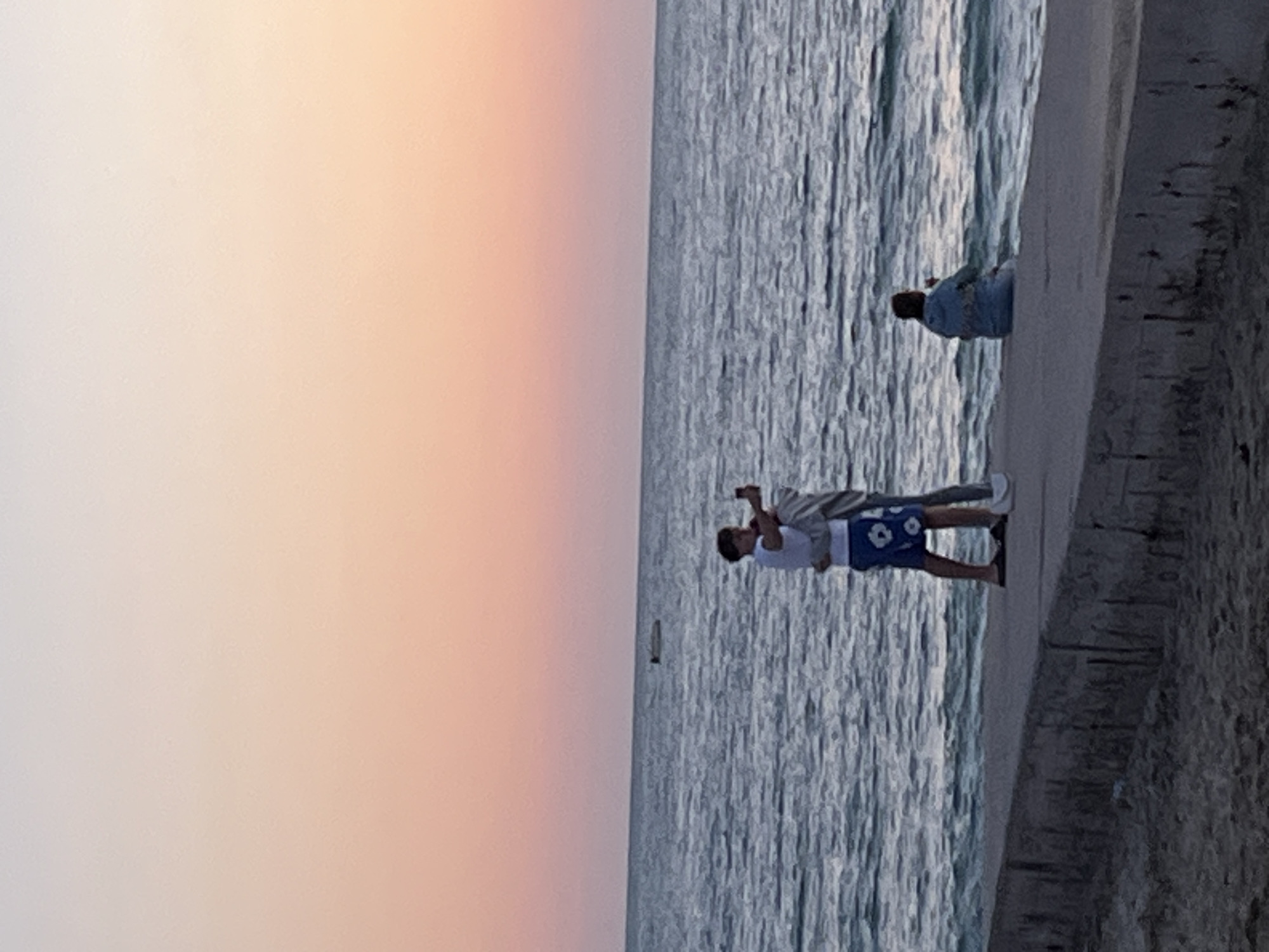 At sunset in Alanya, Turkey, a man in blue floral swim trunks and a white shirt holds a cell phone and takes a picture of a woman. The pair stands on a concrete pier overlooking the water. A third individual sits further down the pier.