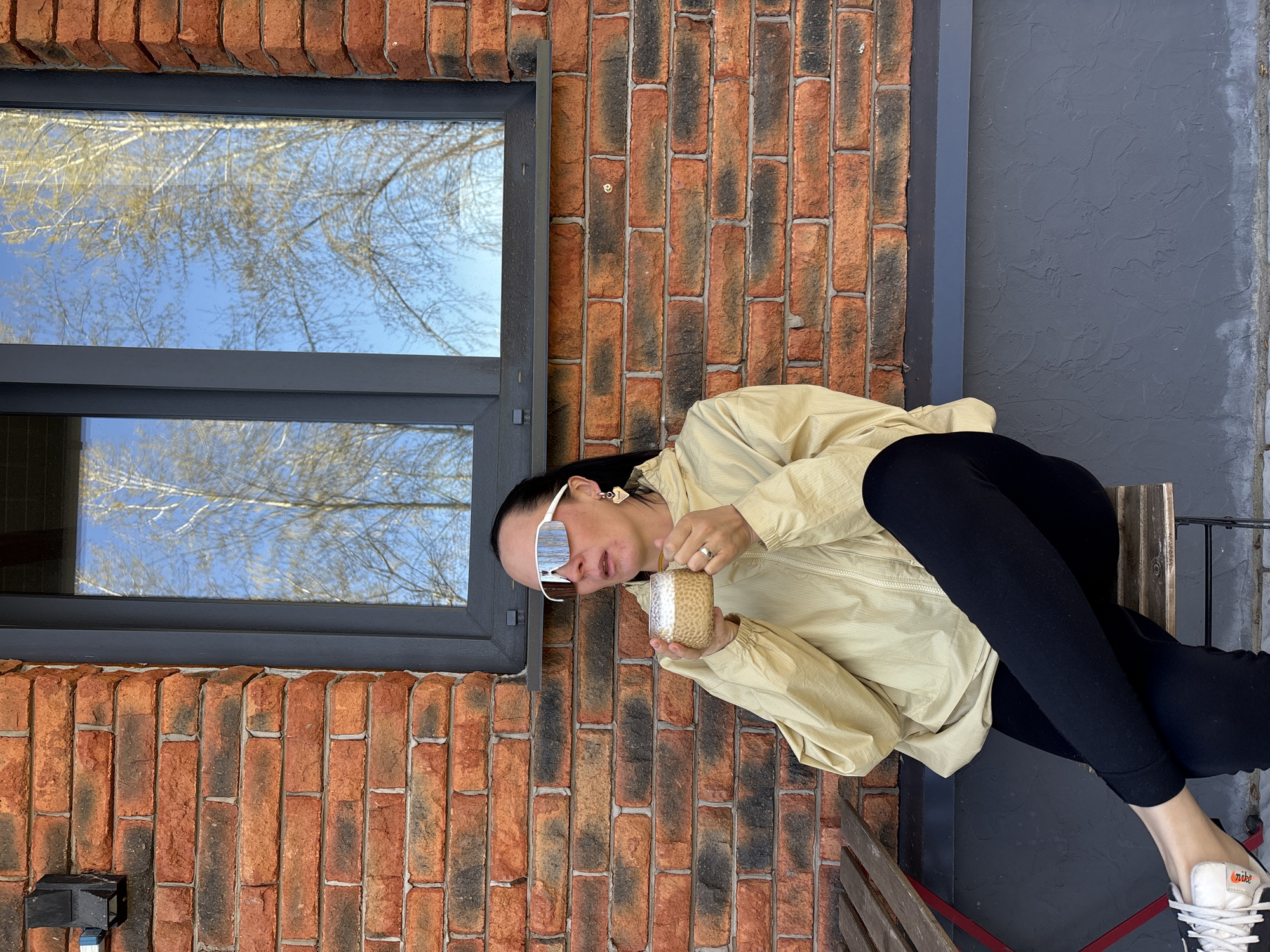 A woman sits on a bench in front of a brick building in Chaikovsky, Russia. The building has a large window reflecting the sky and trees. The woman is wearing sunglasses, a light-colored jacket, and black leggings. She holds a small glass mug. The bench is made of wood and metal. The adjacent wall is gray.