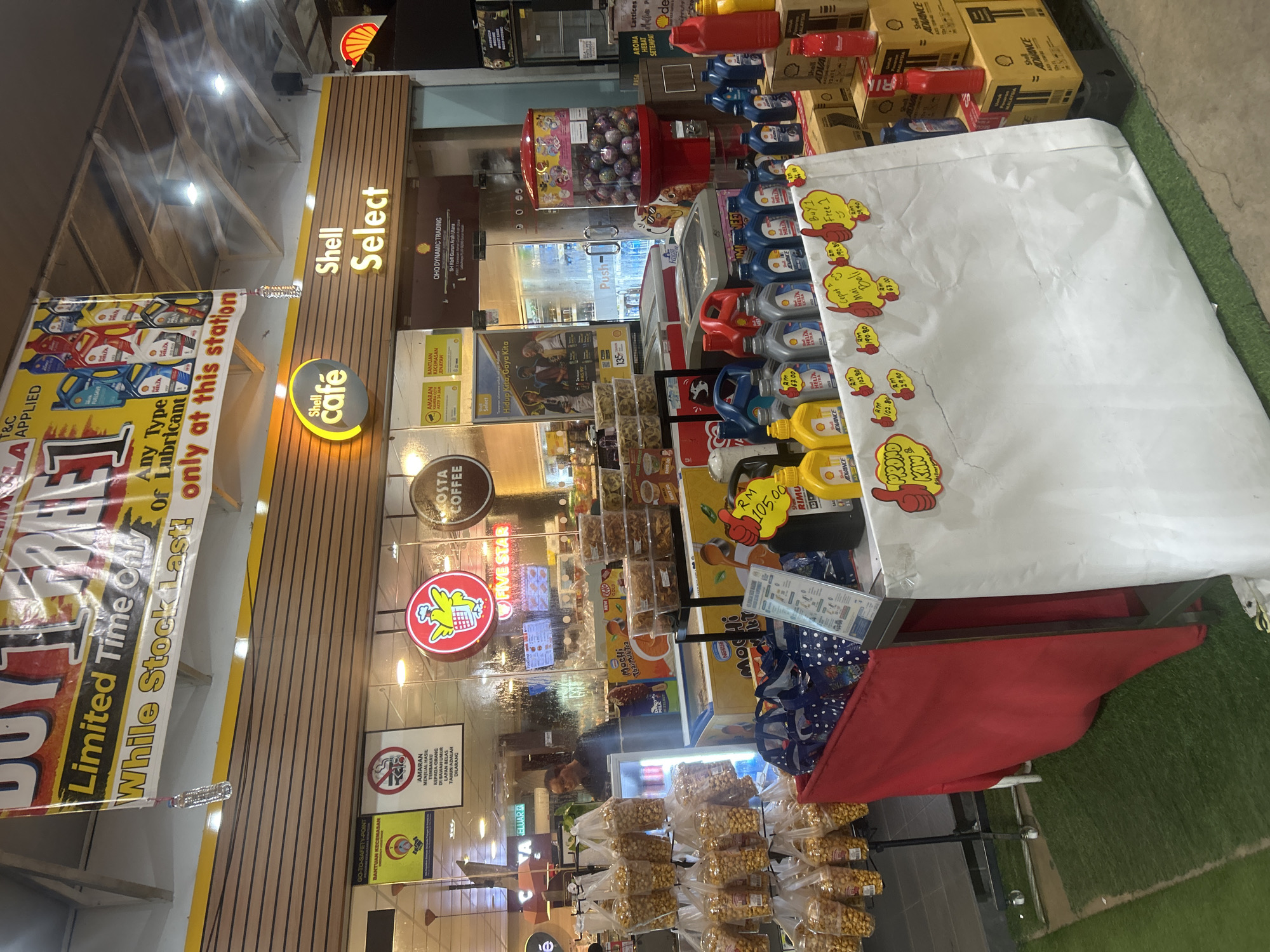 The image depicts the interior of a Shell Select convenience store and a Shell petrol station in Gurun, Malaysia. In the foreground, a promotional display is set up with various engine oil bottles, priced with red tags. A large white sheet with handwritten promotional information and yellow "thumbs up" stickers is draped over a table. Bags of snacks are displayed on shelves to the left, and a red cloth covers a section of the display table. Behind the promotional area, a man is visible inside the store, near a cooler. The Shell Select signage is prominently displayed above the store entrance. The store appears to be part of a larger petrol station complex, as indicated by the Shell branding. The overall environment suggests a retail setting within a petrol station.
