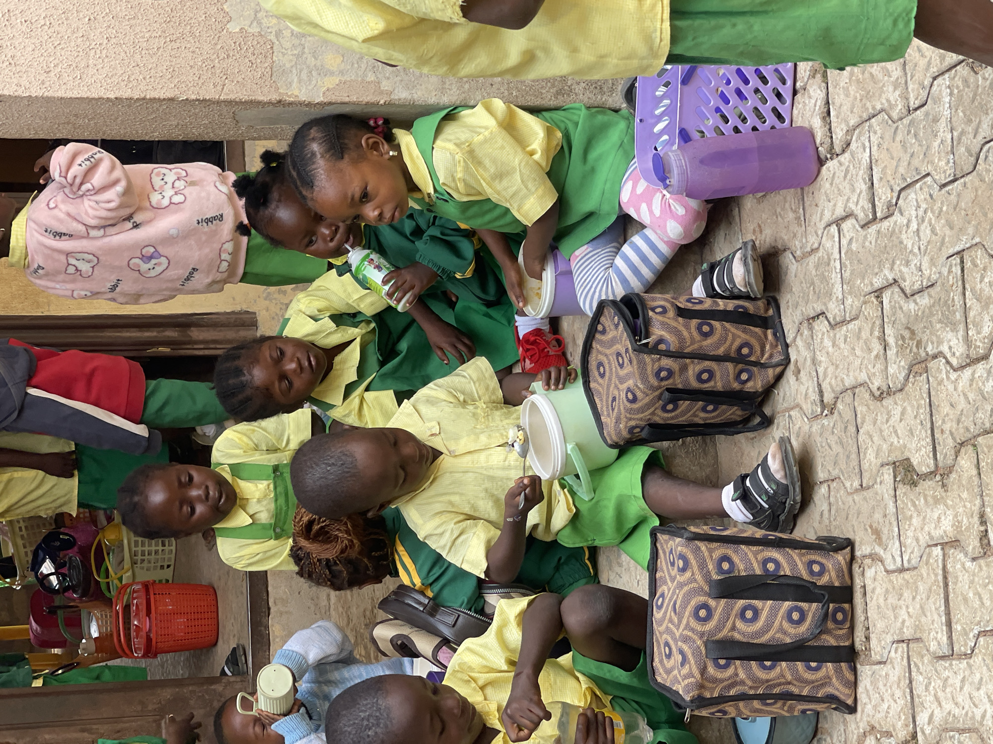 A group of young children, dressed in yellow and green school uniforms, are seated on a textured, light-colored tiled floor outside a building with a tan stucco wall. Some of the children are holding cups and containers, appearing to be eating or drinking. In the foreground, two children are focused on food containers in front of them, one with a spoon digging into a container. To the left, a child is holding a light-colored cup to their mouth. Several patterned lunch bags and a purple water bottle are also visible on the ground. In the background, more children in uniforms can be seen, along with a red basket and other school supplies. The scene suggests a break or mealtime at a school in Akwanga, Nigeria.