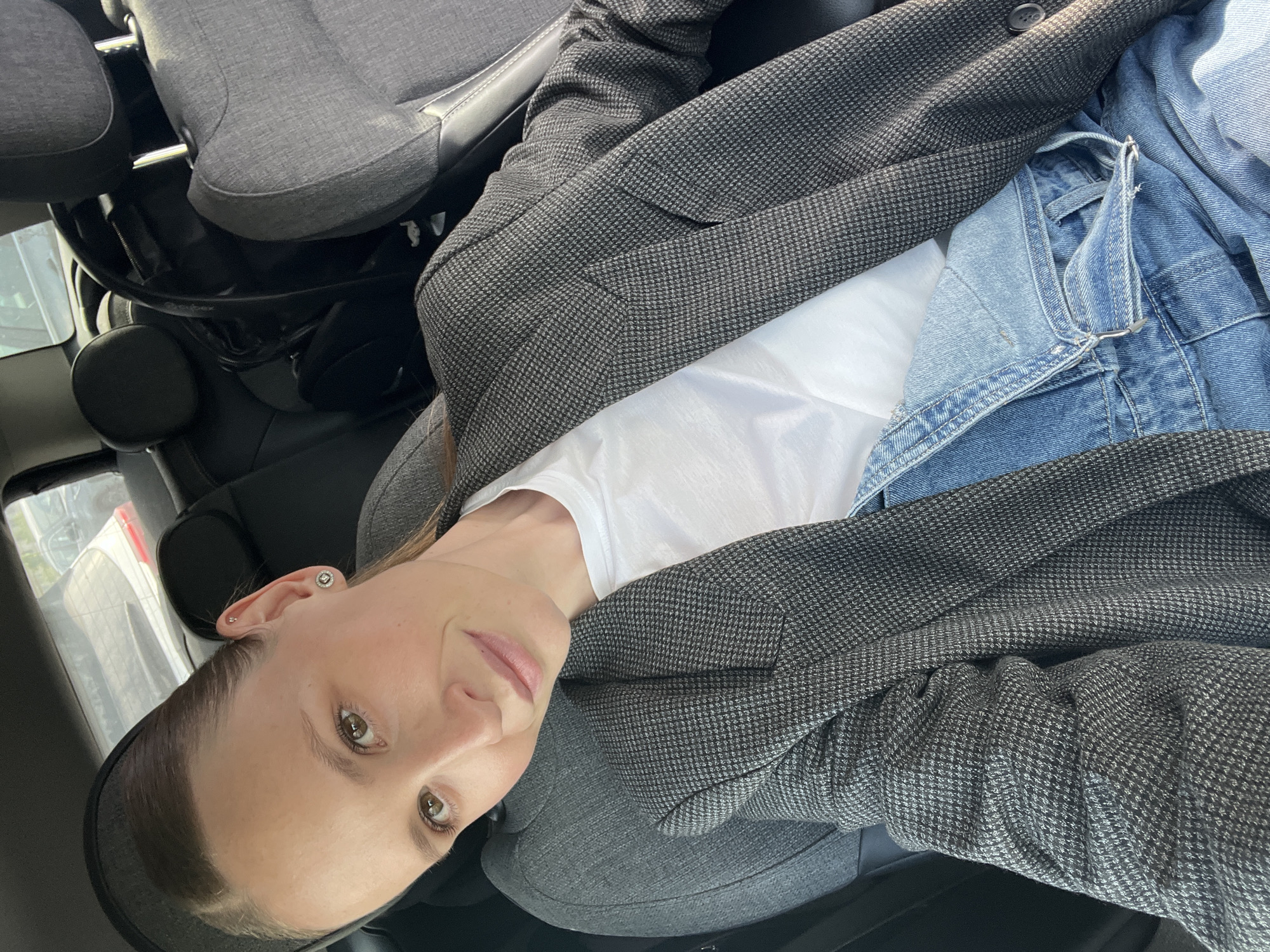 A woman with brown hair and hazel eyes is in the front seat of a car. She is wearing a white t-shirt, blue denim overalls, and a grey houndstooth jacket. To her left is a car seat. The car is parked, as inferred by the presence of parked cars visible through the window. The location is unknown and cannot be determined by this image.