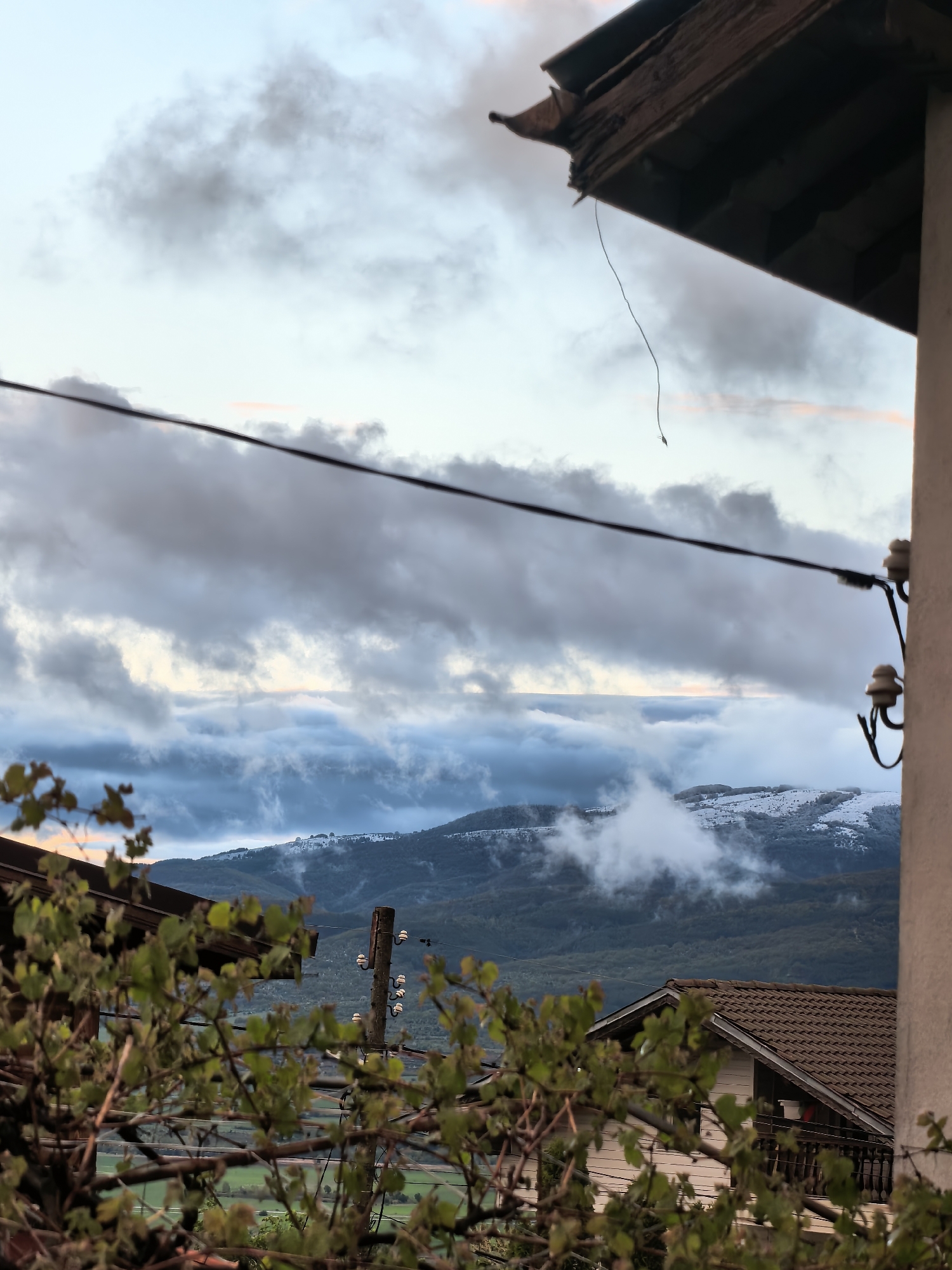This vertical shot captures a mountainous landscape under a cloudy sky, framed by elements of a residential setting, likely in Sapareva Banya, Bulgaria. The time of day appears to be either early morning or late afternoon/dusk, indicated by the soft, diffused light and faint pinkish hues in the upper sky.
The scene is dominated by a dramatic cloudscape. Dark, heavy clouds fill the mid-sky, while lower, lighter clouds cling to and obscure the distant mountains. Patches of snow are visible on the mountain peaks, suggesting a cold season or high altitude. The mountains themselves are covered in dense forest.
In the immediate foreground, dark, blurred green foliage, possibly vines or a bush, frames the bottom and left side of the image. Architectural elements partially frame the view: the weathered, wooden underside of a roof overhang with a hanging string is visible in the top right, and a light-colored vertical wall with two ceramic insulators for utility lines is present on the far right. A prominent dark utility wire stretches horizontally across the mid-section of the image.
Further into the midground, a house with a brown tiled roof and a balcony is partially visible on the bottom right, behind the foliage. To its left, a wooden utility pole with multiple ceramic insulators stands. No human activity or visible text is present. The overall mood is serene and somewhat moody due to the cloudy weather and soft light.