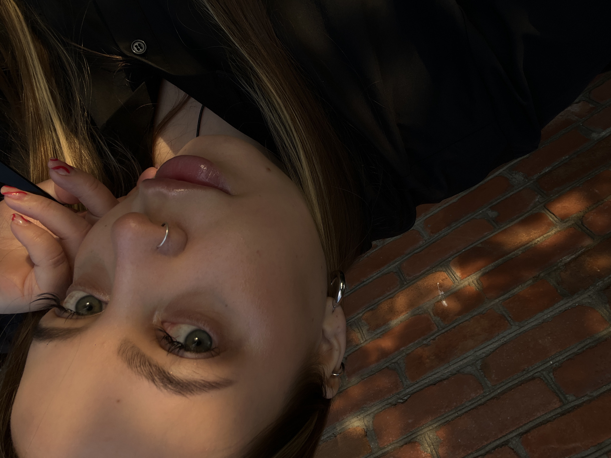 A close-up, upward-angled portrait features a young individual, likely female, against a brick wall. The subject's face is prominent, tilted with the crown of the head towards the upper right. They have light-colored (green/hazel) eyes and long brown hair with blonde highlights. A silver hoop nose ring is visible on their left nostril, and multiple silver ear piercings, including a helix cuff and a lobe ring, adorn their right ear. Their lips are slightly parted and appear glossy. One hand, displaying red and white patterned nail polish, rests near their left cheek. The subject wears dark, possibly black, clothing with a dark button detail visible on a collar or lapel.
The background is a reddish-brown brick wall with grey mortar. Strong, directional lighting from the upper left casts distinct shadows across the subject's face and creates patterns of light and shadow on the brickwork. The individual's gaze is directed upwards towards the camera, conveying a serene or contemplative expression. No text is visible in the image. The scene is located in Novopokrovskaya, Russia.