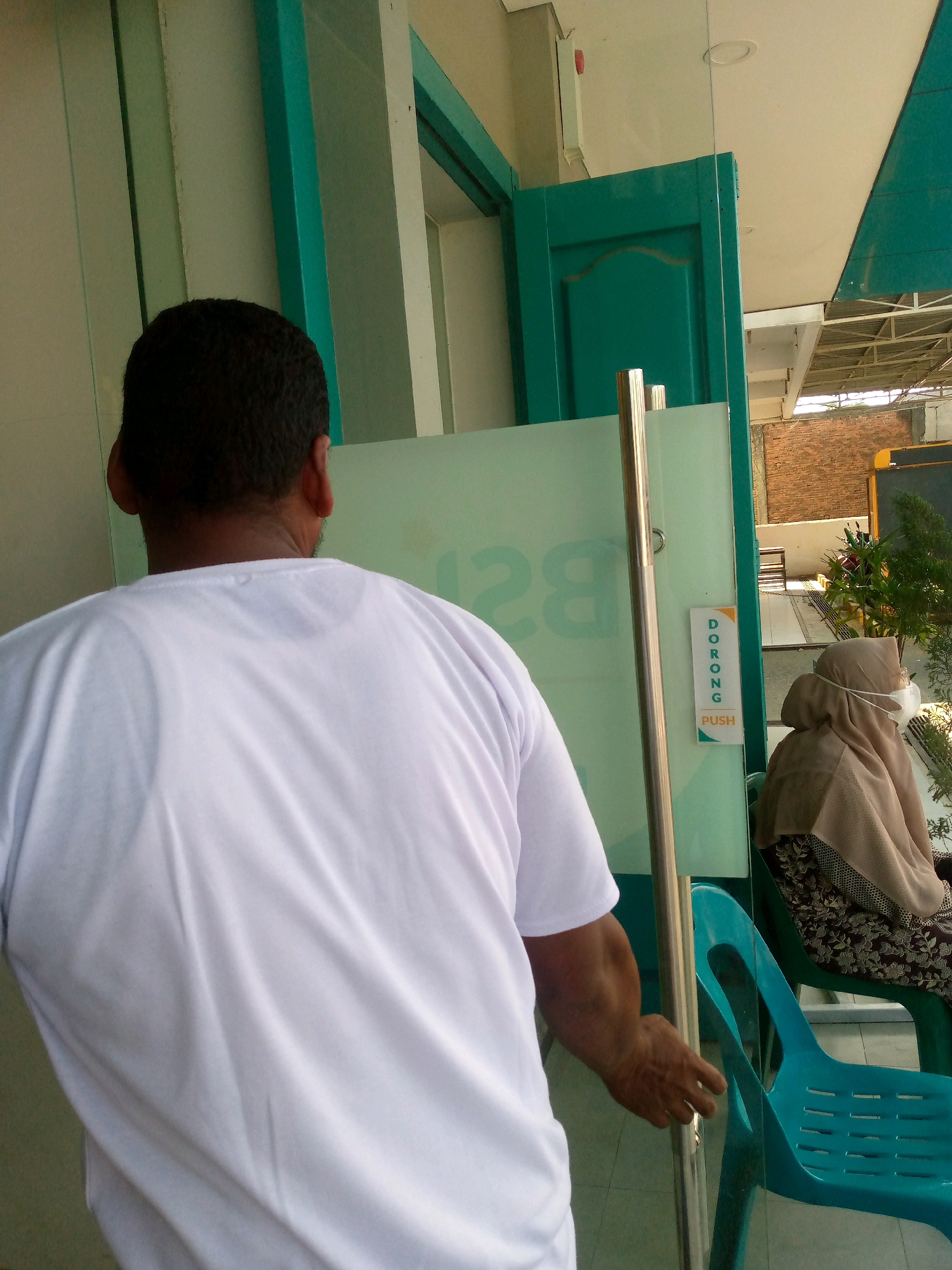 The image captures an outdoor scene in Blangpeuria, Indonesia, with a focus on a man from the chest up, wearing a white t-shirt. He is positioned in the foreground, seen from behind, with his right arm extended towards a glass door. His hand appears to be in the process of pushing the door open. The glass door has a vertical metal handle and a small sign that reads "DORONG PUSH" in orange and white lettering. Behind the man and the door, a woman wearing a beige hijab and a face mask is visible, seated on a teal plastic chair. She is partially obscured by the glass door. The environment appears to be an entrance to a building, with a green wooden doorframe visible to the left and a ceiling with exposed structure to the right, suggesting an open-air or semi-enclosed area. The background reveals a brick wall and foliage. The lighting indicates it is daytime.
