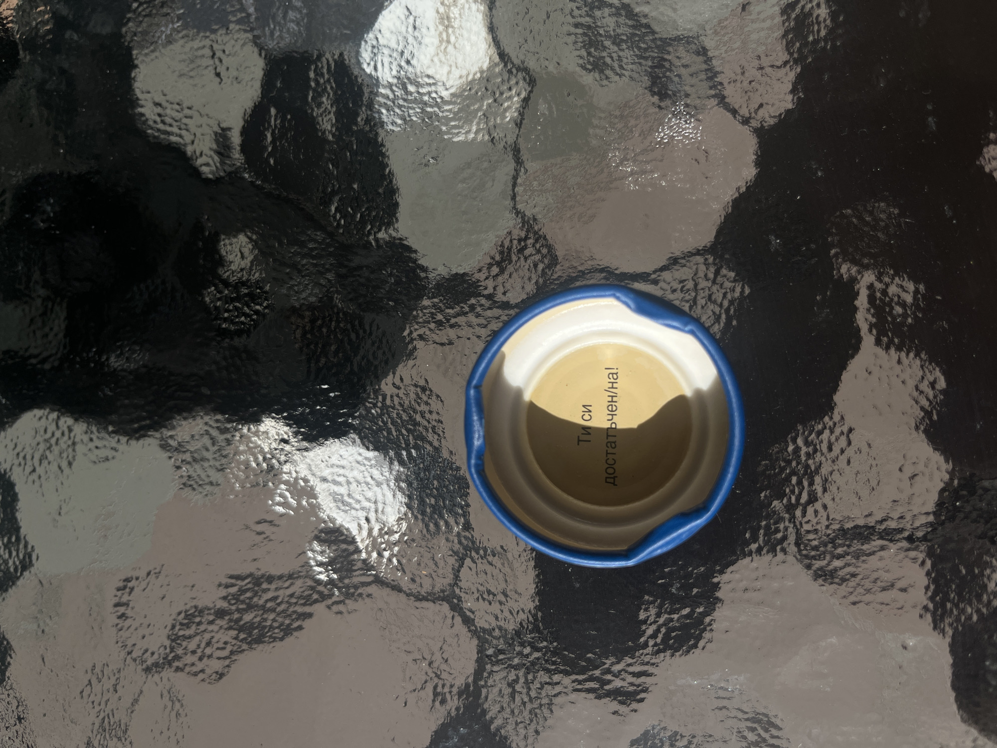 The image captures a top-down, close-up view of a blue and white cap resting on a dark, highly textured, and reflective surface, likely patterned glass, such as a patio table. The surface shows irregular ripples and distortions, reflecting bright sunlight and darker shadows, indicating an outdoor setting on a clear, sunny day. Inside the cap, which has a light, off-white interior, there is visible text in Cyrillic script. The text reads, oriented vertically, "Ти си достатъчен/на!". This phrase translates from Bulgarian to "You are enough!" or "You are sufficient!", with the "ен/на" indicating gender agreement for the adjective. No people or specific activities are depicted, focusing instead on this singular object and its reflective environment. The strong contrasts of light and shadow suggest direct sunlight.