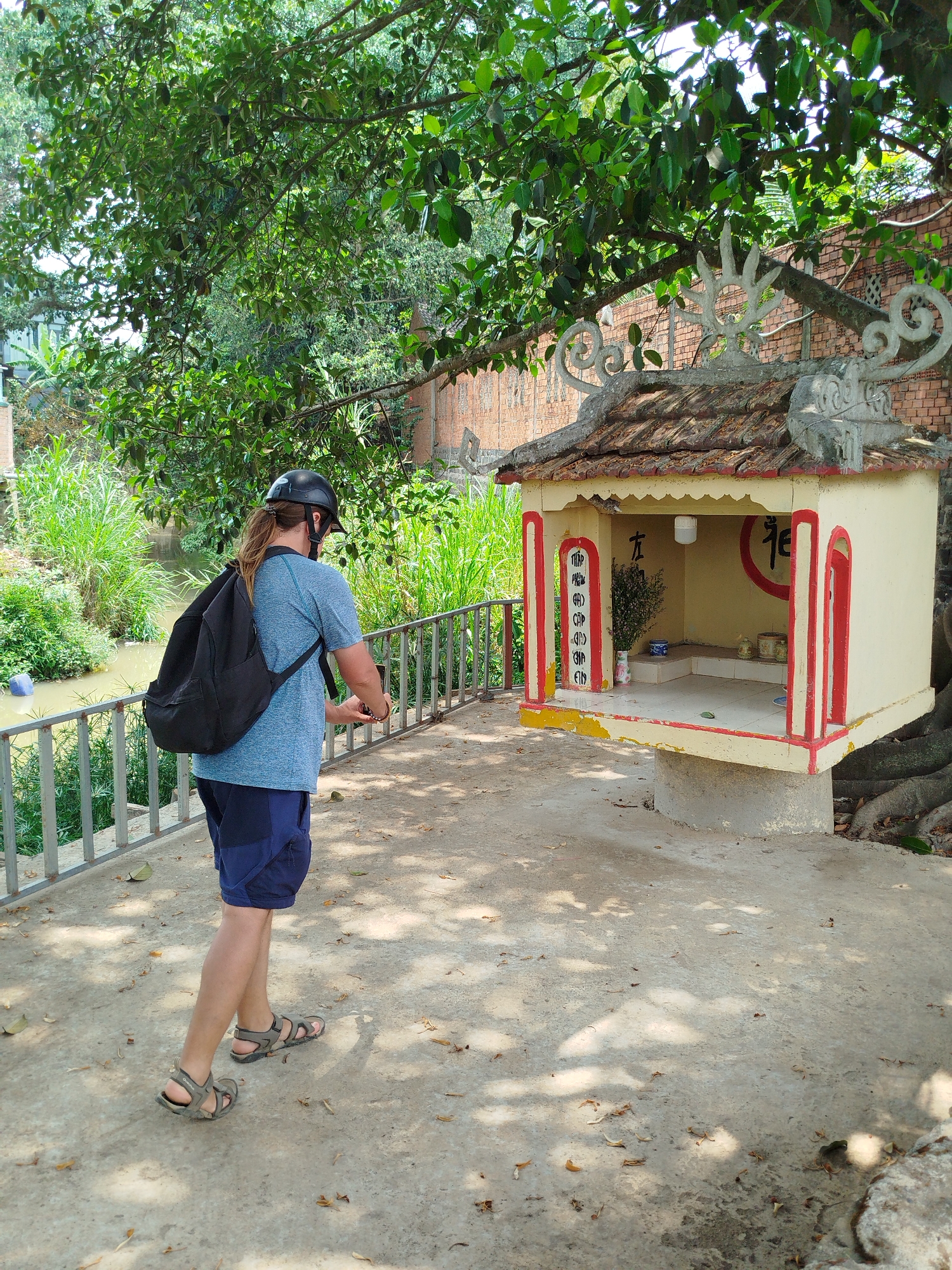 A full-length shot shows a person from the rear, standing on a concrete slab near a small, decorated structure, potentially in Phú Thạnh, Vietnam. The person wears a black helmet, a blue t-shirt, dark blue shorts, and hiking sandals. A black backpack is slung over their shoulder. They are holding a phone. To the left, a metal railing separates the concrete slab from a green, watery area with dense vegetation. The small structure is a beige cuboid with a tiled roof, red trim, and ornate decorations on the roofline. Inside, there are small vases, a circular symbol and Chinese text on the walls. The structure appears to be a shrine. Overhanging trees provide shade.
