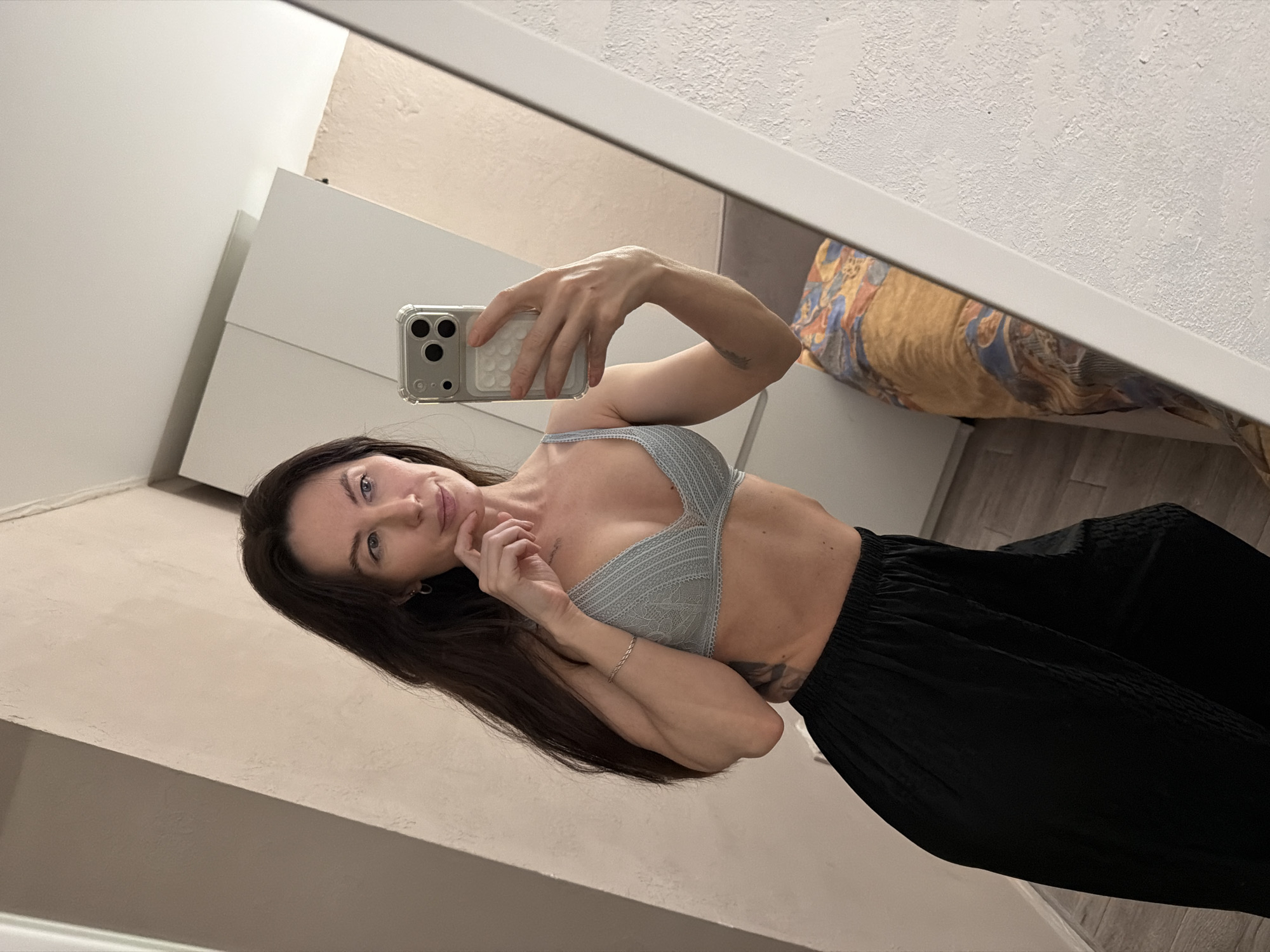 A woman is taking a selfie in a mirror. She is wearing a light blue bralette and black pants. She is holding a phone with a clear case. The background includes a white wall, a white dresser, and a bed with a patterned comforter. The photo appears to be taken indoors. There is no verifiable indication that the location is in Perm, Russia.