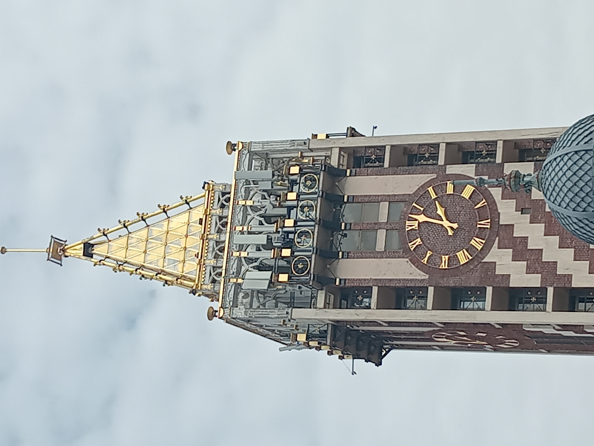 The image shows a close-up, low-angle view of the Astronomical Clock tower in Batumi, Georgia. The clock tower features a gold-colored spire, a complex mechanical structure below the spire, and a large clock face with Roman numerals. The clock face shows the time as approximately 9:53. The building facade is decorated with a brown and white zigzag pattern. The sky is overcast.