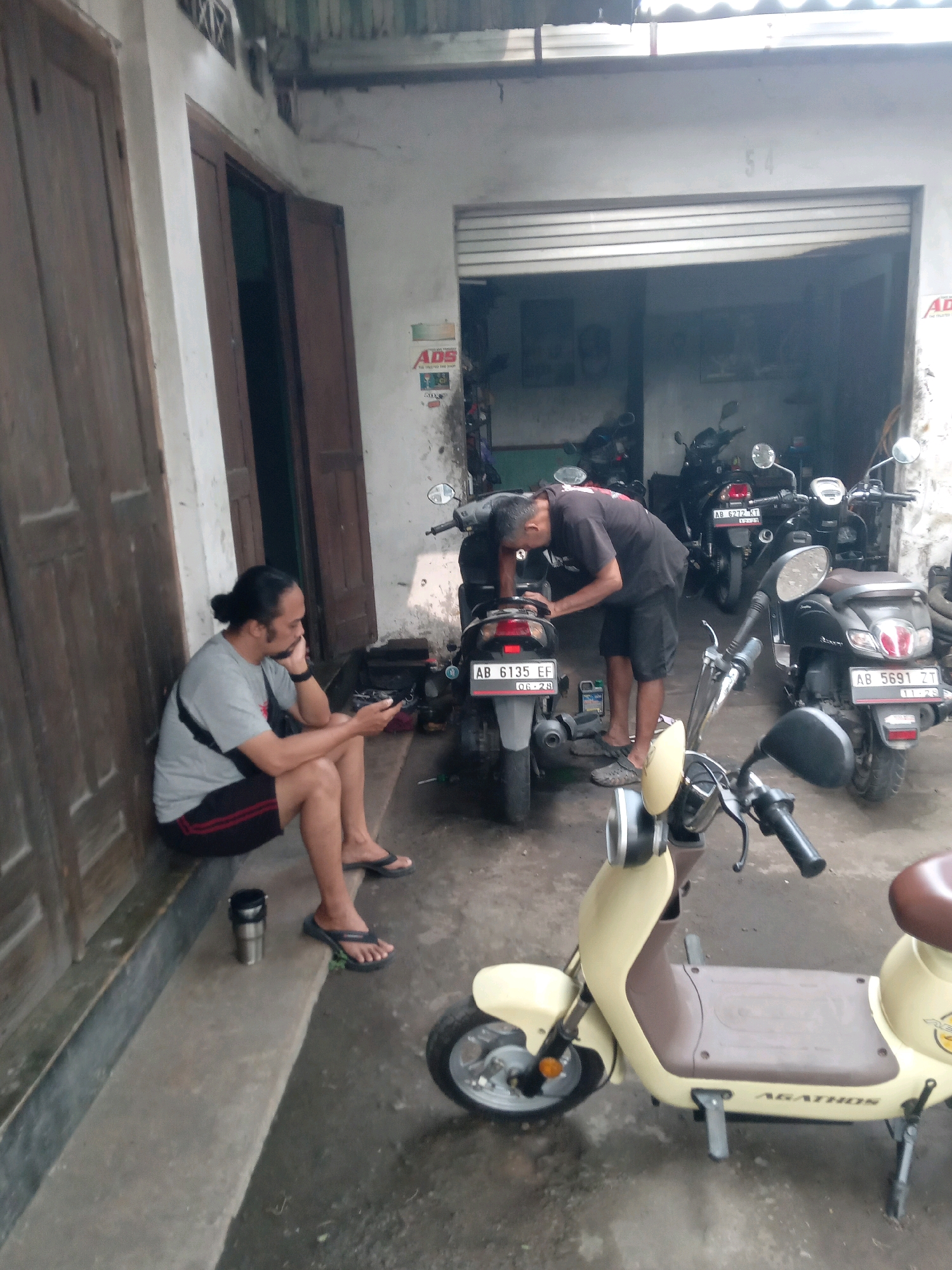 In Banguntapan, Indonesia, an outdoor scene depicts two men near a garage or workshop. On the left, a man with his hair tied back sits on a ledge, looking at his phone. He wears a grey t-shirt, black shorts with red stripes, and black flip-flops. A black bag is slung across his chest. Next to him, a metallic thermos sits on the ledge. In the center of the image, a second man, wearing a black t-shirt and black shorts, is bent over a dark-colored scooter, appearing to be working on it. The scooter has a license plate reading "AB 6135 EF". Several other scooters are visible in the background, some parked inside the garage and others outside. One scooter in the foreground is light yellow and brown, with "AGATHOS" printed on its side. The environment appears to be a street or a workshop area with a concrete floor, some of which is wet. A large wooden door is visible on the left side of the frame, and a corrugated metal overhang is above the garage entrance. A sign with "ADS" is affixed to the wall near the garage.