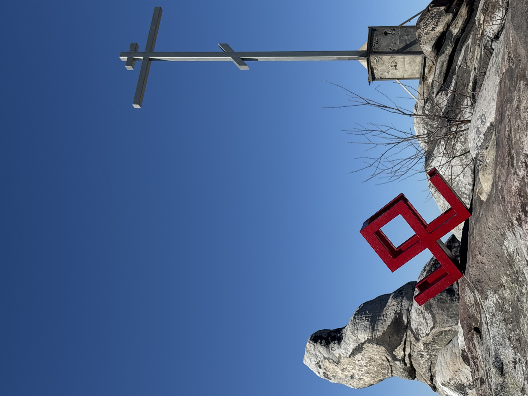 The image presents an upward shot of a rocky terrain against a clear blue sky. Two significant structures are prominent: a tall, simple, gray Christian cross on a concrete base is positioned at the top of the frame. A red geometric artwork is installed on the side of the rock formation. The photo was taken in Gubakha, Russia.